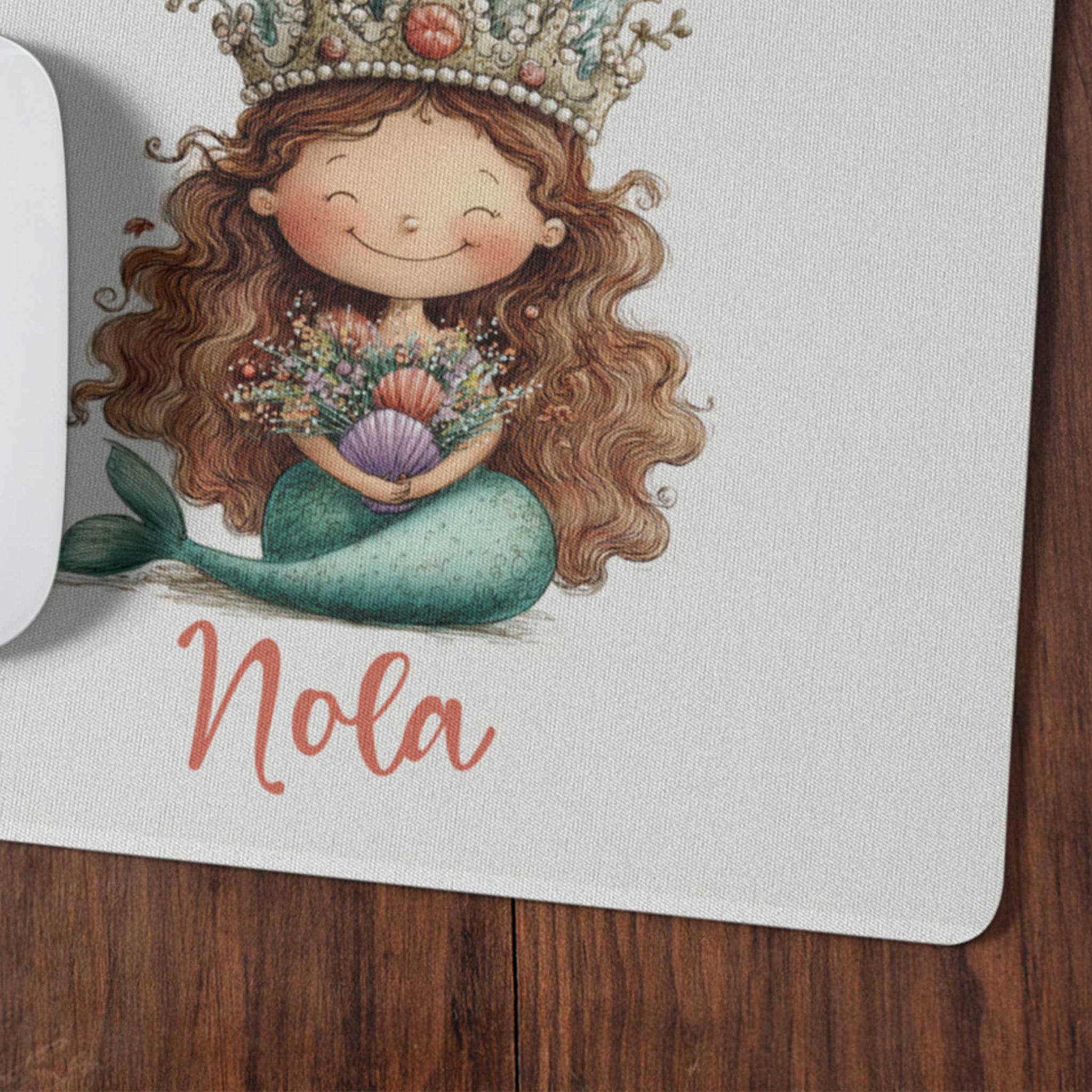 Close-up van een witte muismat met een zeemeermin met lange bruine haren, een koraalkroon en de naam Nola op een houten bureau.