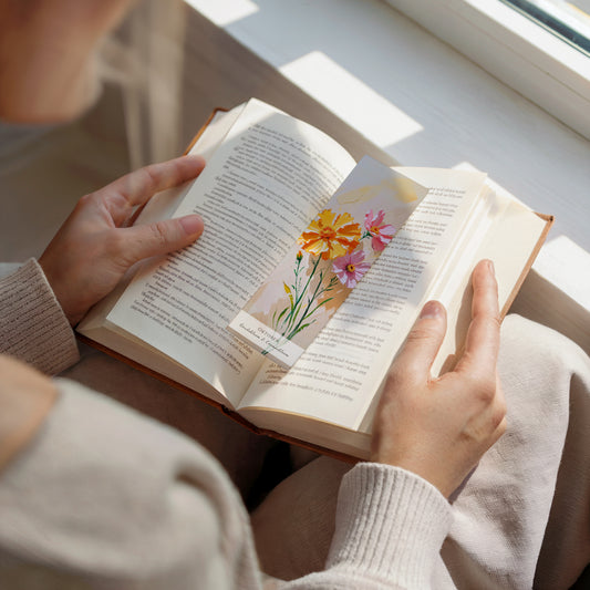 Een vrouw die een boek leest bij het raam. Op het open boek ligt een bladwijzer. Met een geboorte bloem boekenlegger voor de maand oktober, met  kunstzinnige illustratie van Goudsbloem & Cosmosbloem.