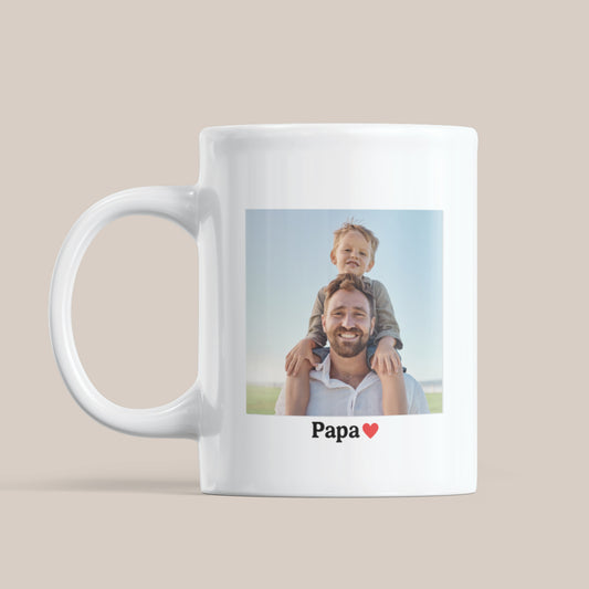 Witte keramische mok op een beige achtergrond met een foto van een vader met kind op zijn schouders en daaronder de tekst “Papa” met een rood hartje.