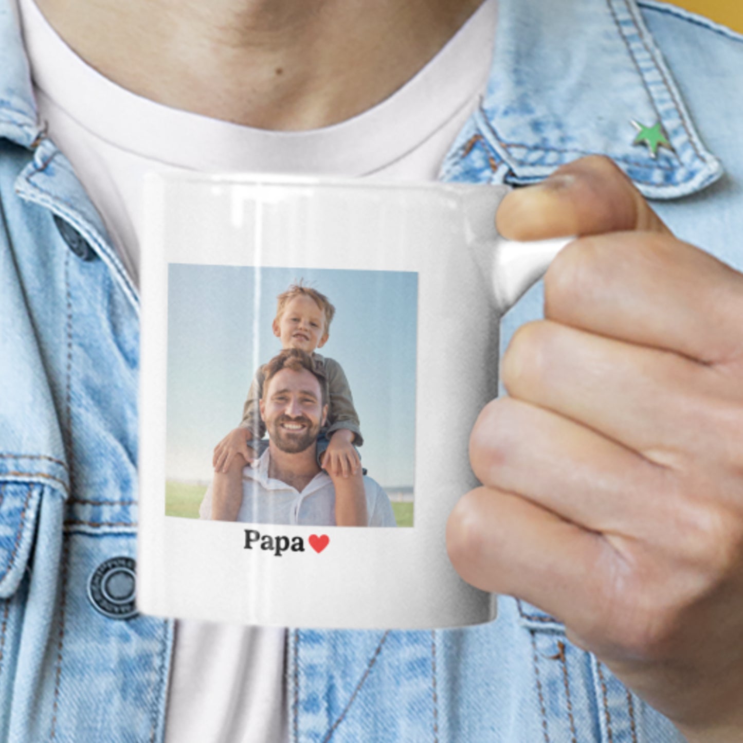 Close-up van een witte keramische mok die wordt vastgehouden, met daarop een foto van een vader met kind op zijn schouders en de tekst “Papa” met een rood hartje.