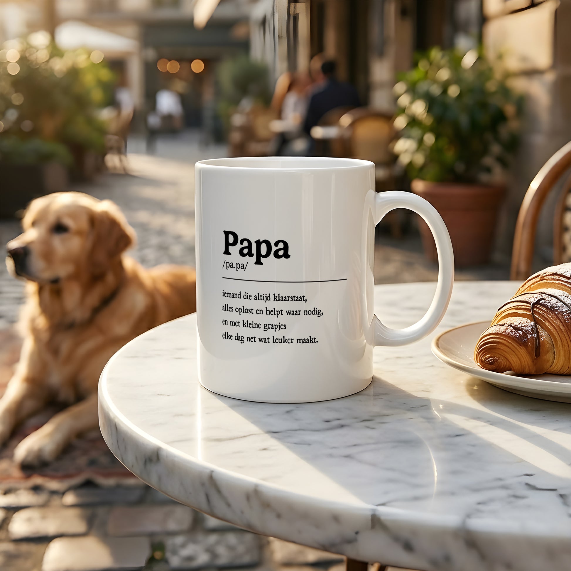Witte mok met papa tekst op een tafel in warme buiten setting met croissant en hond op de achtergrond.