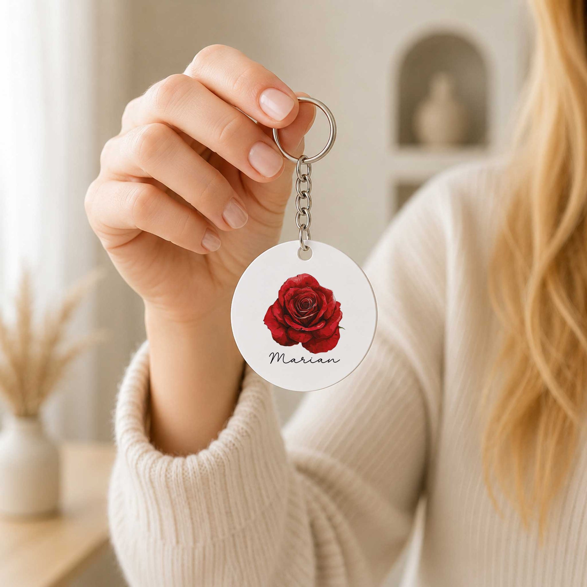 Vrouw houdt een ronde sleutelhanger met een rode roos en de naam Marian vast aan een metalen sleutelring in een lichte woonkamer.