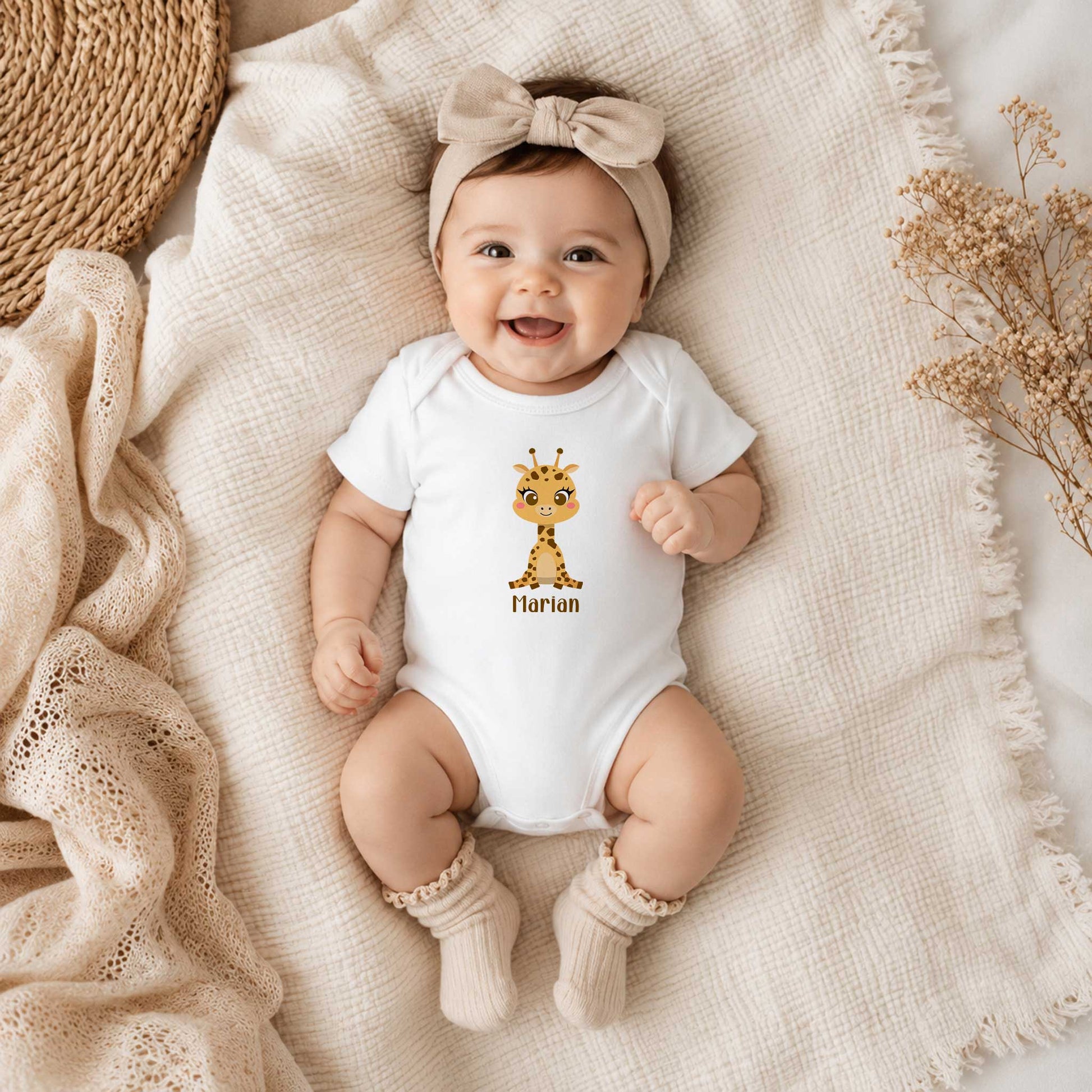Lachende baby draagt een witte romper met een schattig girafje en de naam Marian en ligt op een beige hydrofiele deken met een strikhaarband.