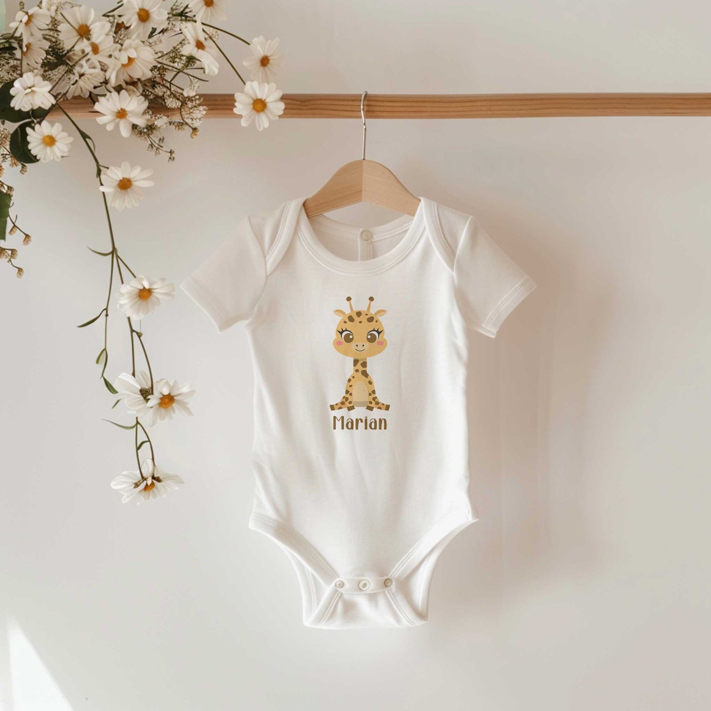 Witte babyromper met een schattig girafje en de naam Marian hangt aan een houten kledinghanger met madeliefjes op de achtergrond.