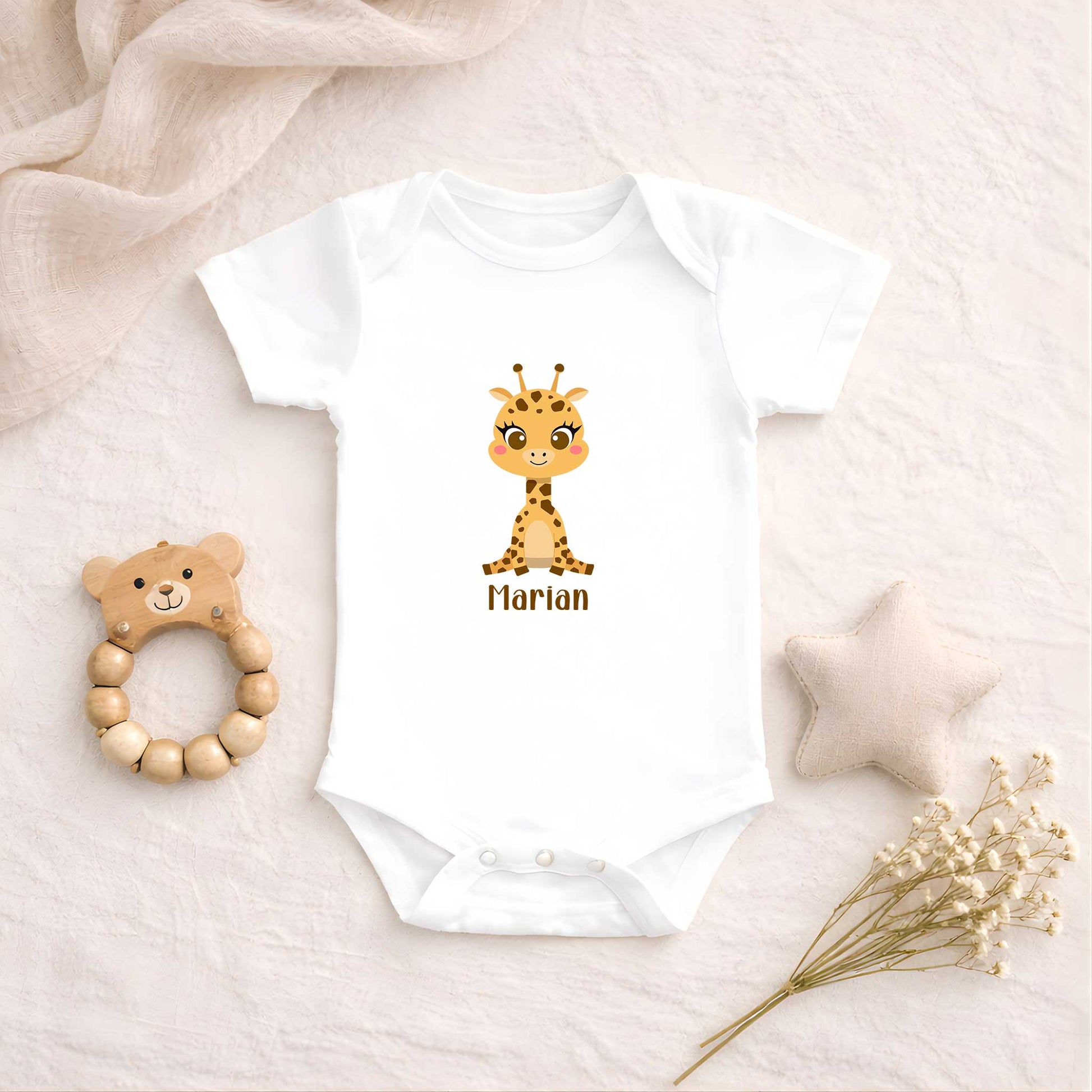Witte babyromper met een schattig girafje en de naam Marian ligt op een zachte beige ondergrond naast een houten bijtring, een stoffen ster en droogbloemen.