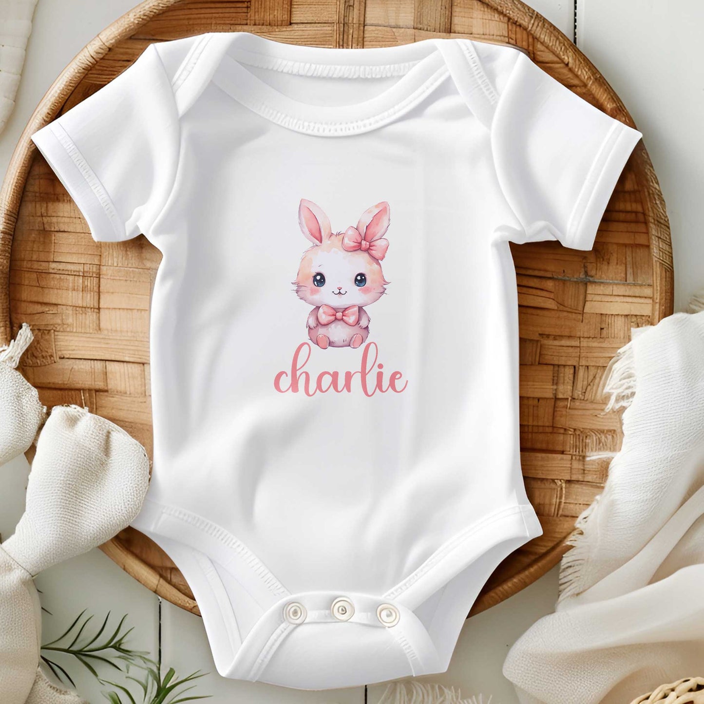 Witte romper met kawaii konijntje en roze strikje, bedrukt met de naam ‘charlie’ in sierlijk roze lettertype, gefotografeerd op een bamboe schaal.