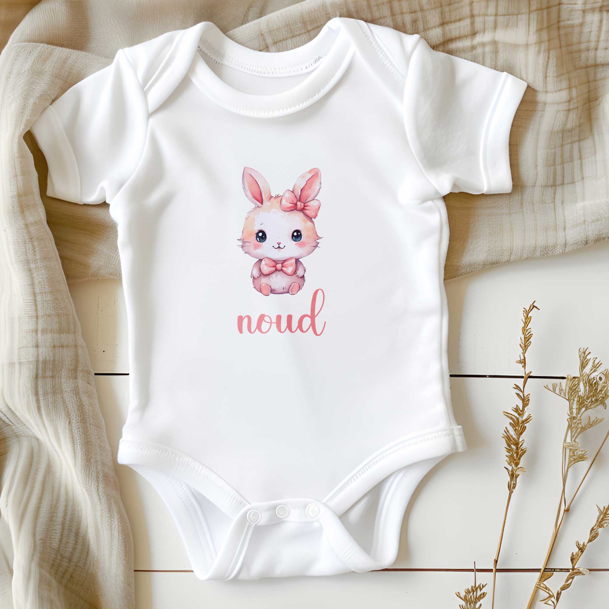 Wit babyrompertje op een houten ondergrond met daarop een kawaii konijntje met roze strik en de naam ‘noud’ in roze tekst.