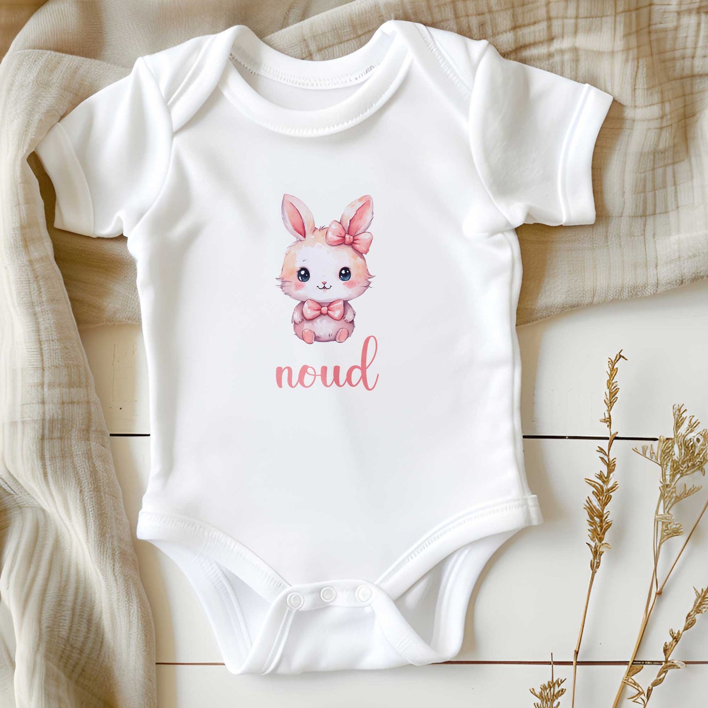 Wit babyrompertje op een houten ondergrond met daarop een kawaii konijntje met roze strik en de naam ‘noud’ in roze tekst.