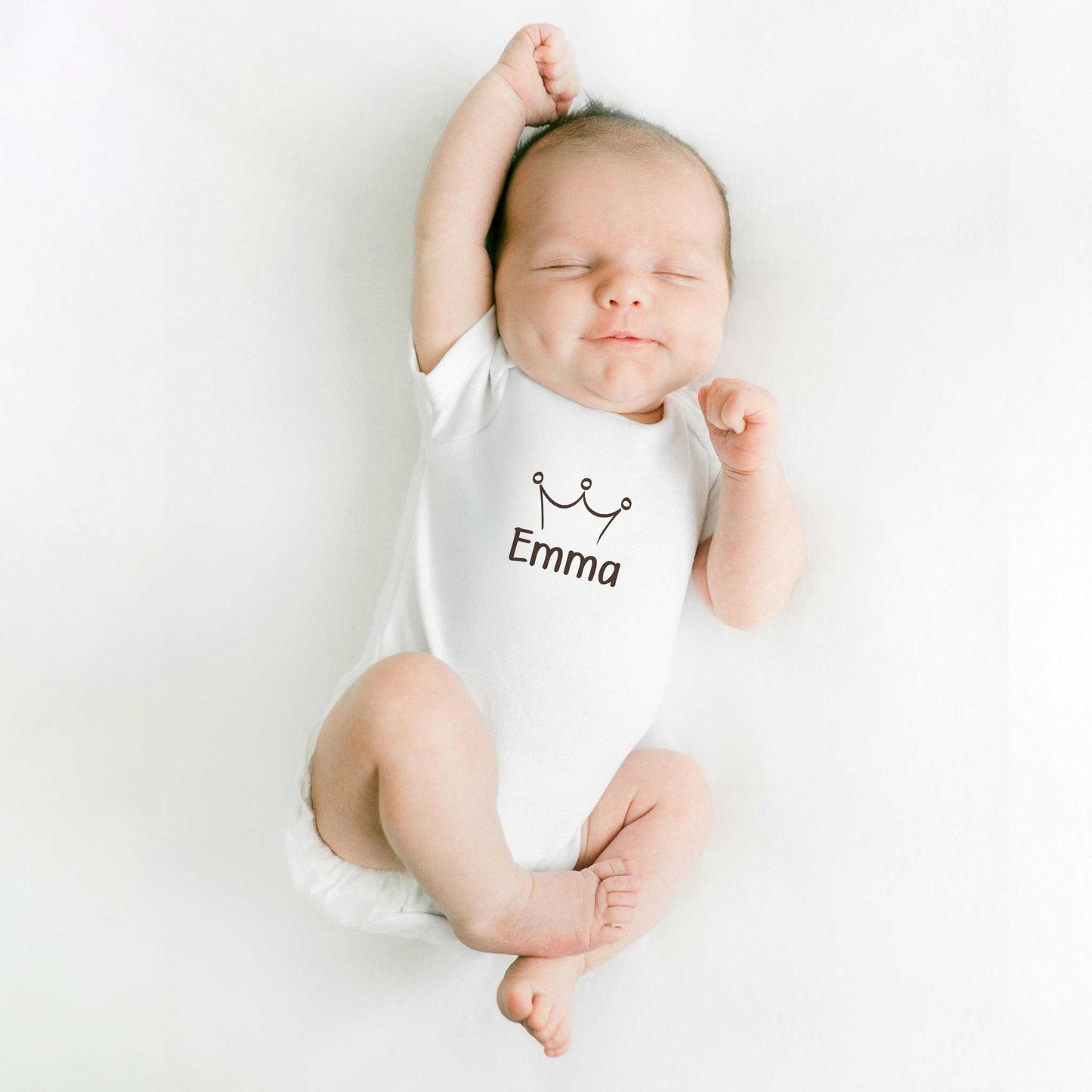 Slapende baby draagt een witte romper met een kroontje en de naam Emma en ligt op een witte ondergrond met één arm omhoog.