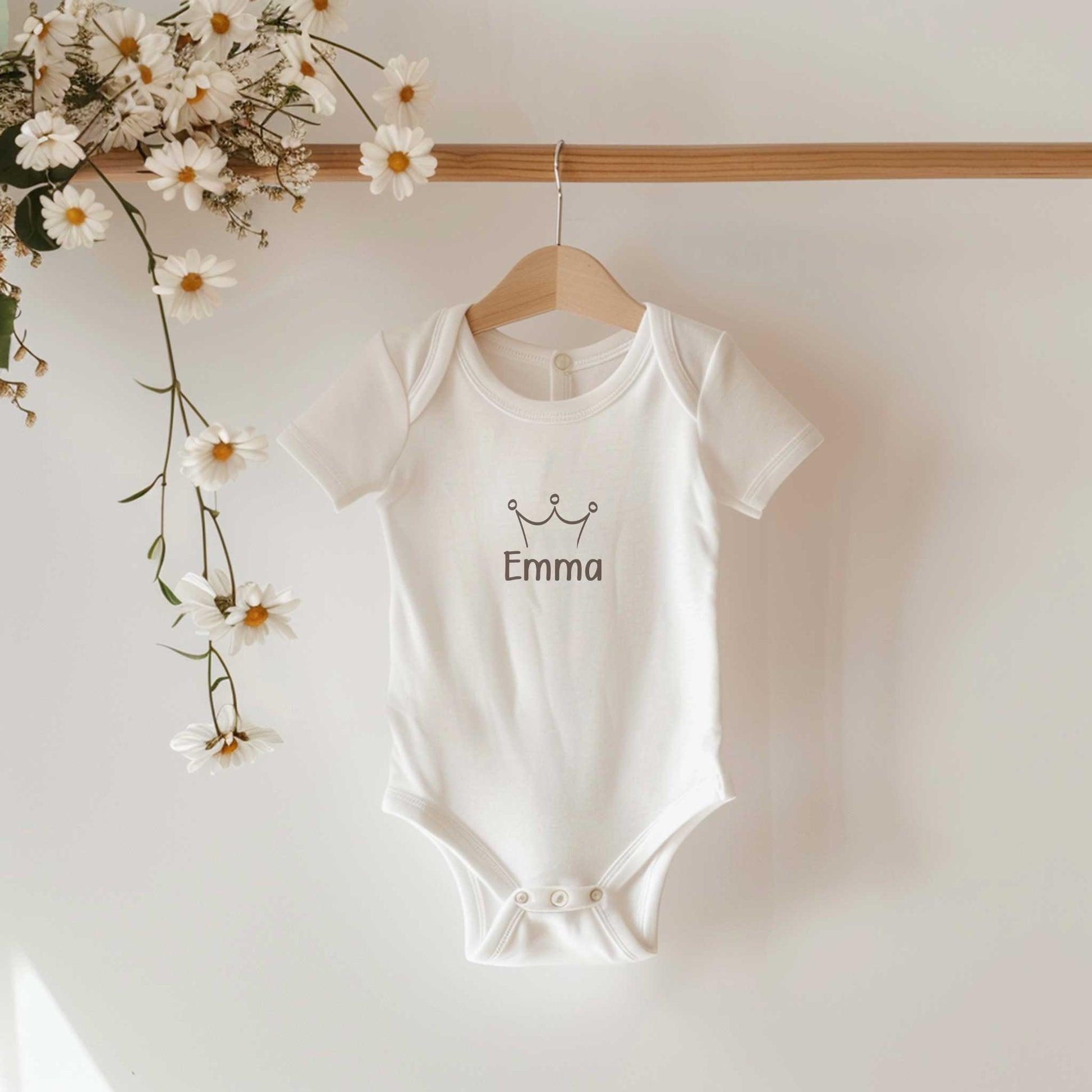 Witte babyromper met een kroontje en de naam Emma hangt aan een houten kledinghanger met madeliefjes op de achtergrond.