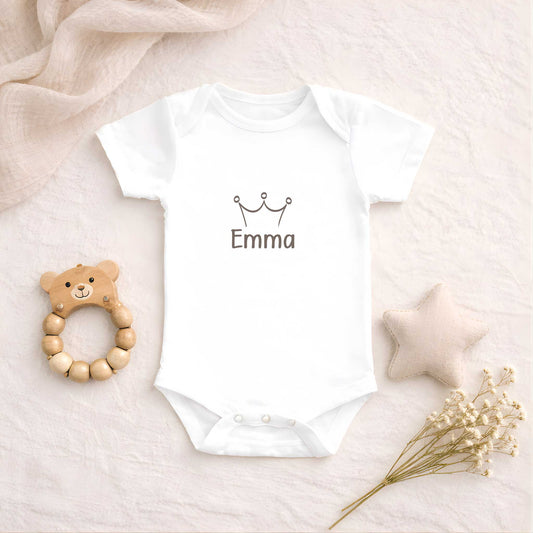 Witte babyromper met een kroontje en de naam Emma ligt op een zachte beige ondergrond naast een houten bijtring, een stoffen ster en droogbloemen.