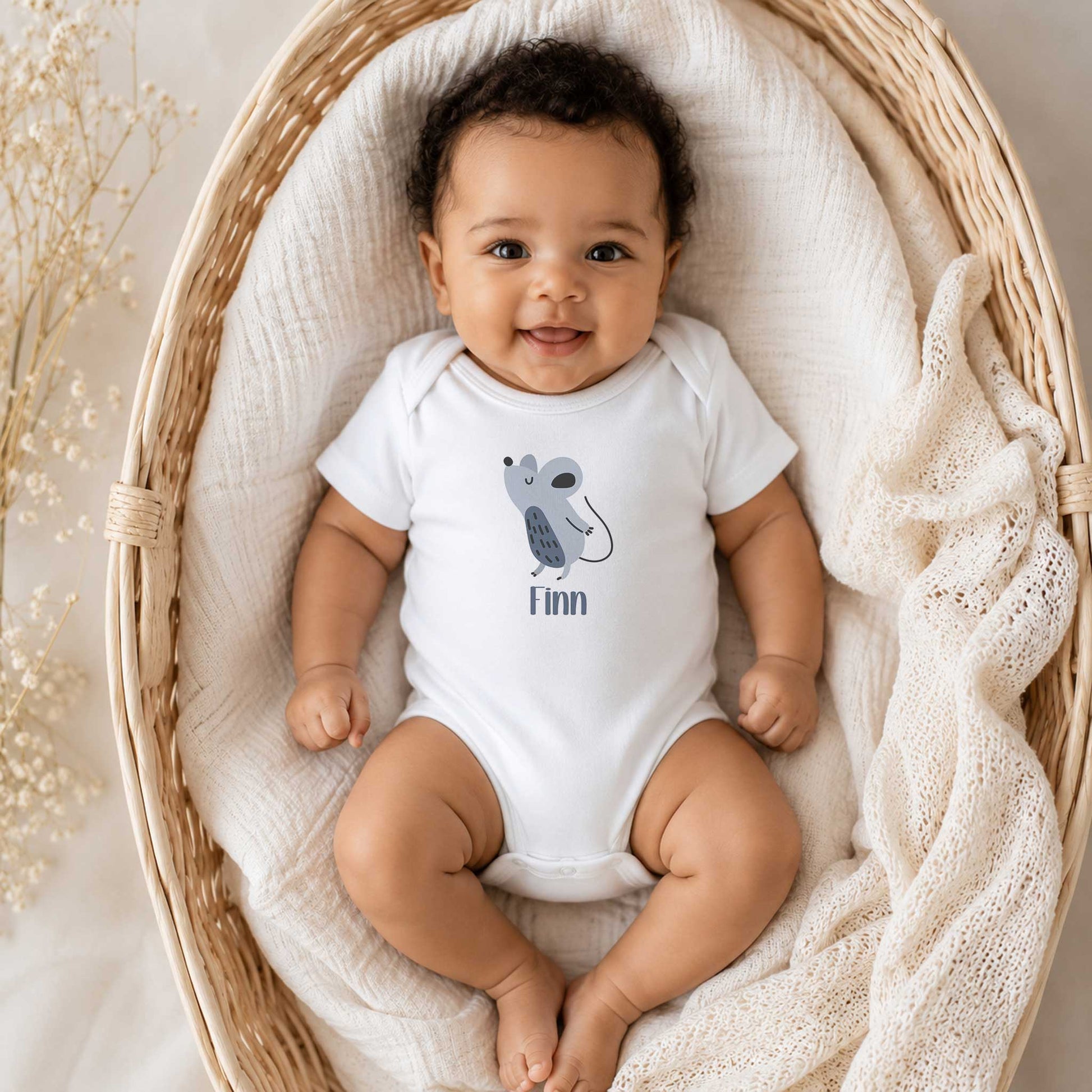 Lachende baby ligt in een rotan babymand en draagt een witte romper met een schattige muis en de naam Finn.
