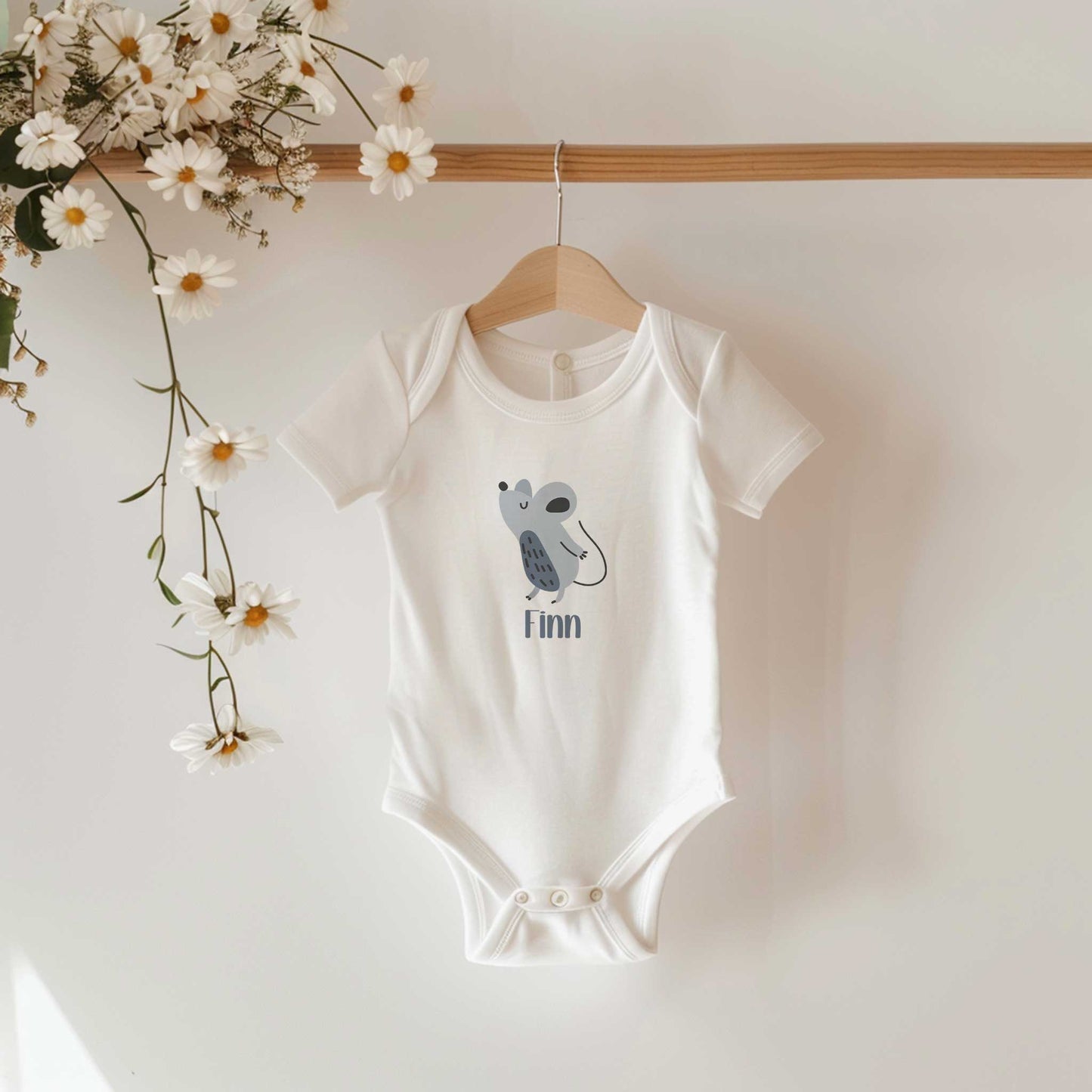 Witte romper met een schattige muis en de naam Finn hangt aan een houten kledinghanger naast witte bloemen.