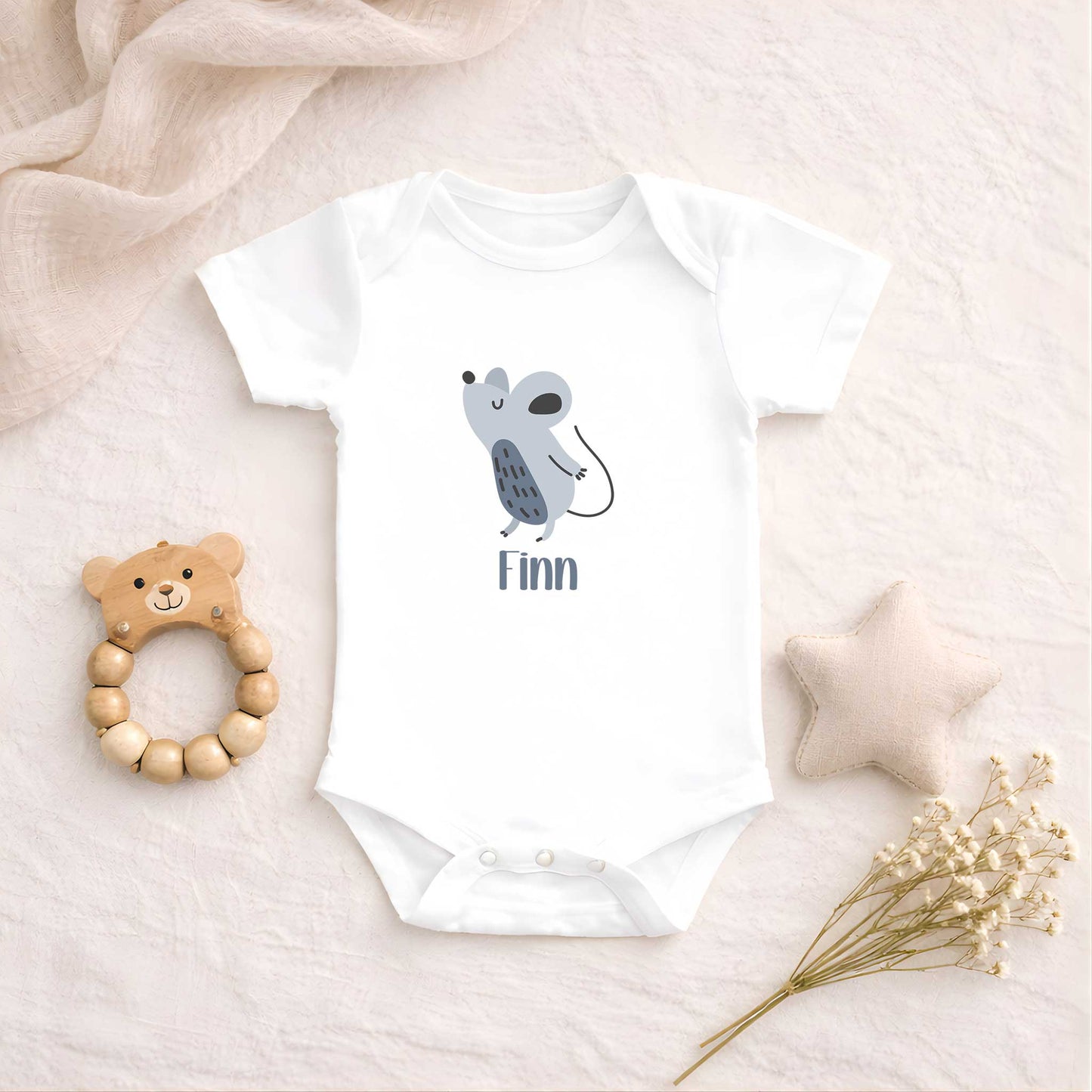 Witte romper met een schattige muis en de naam Finn ligt op een zachte beige ondergrond naast een houten bijtring, stervormig kussen en droogbloemen.