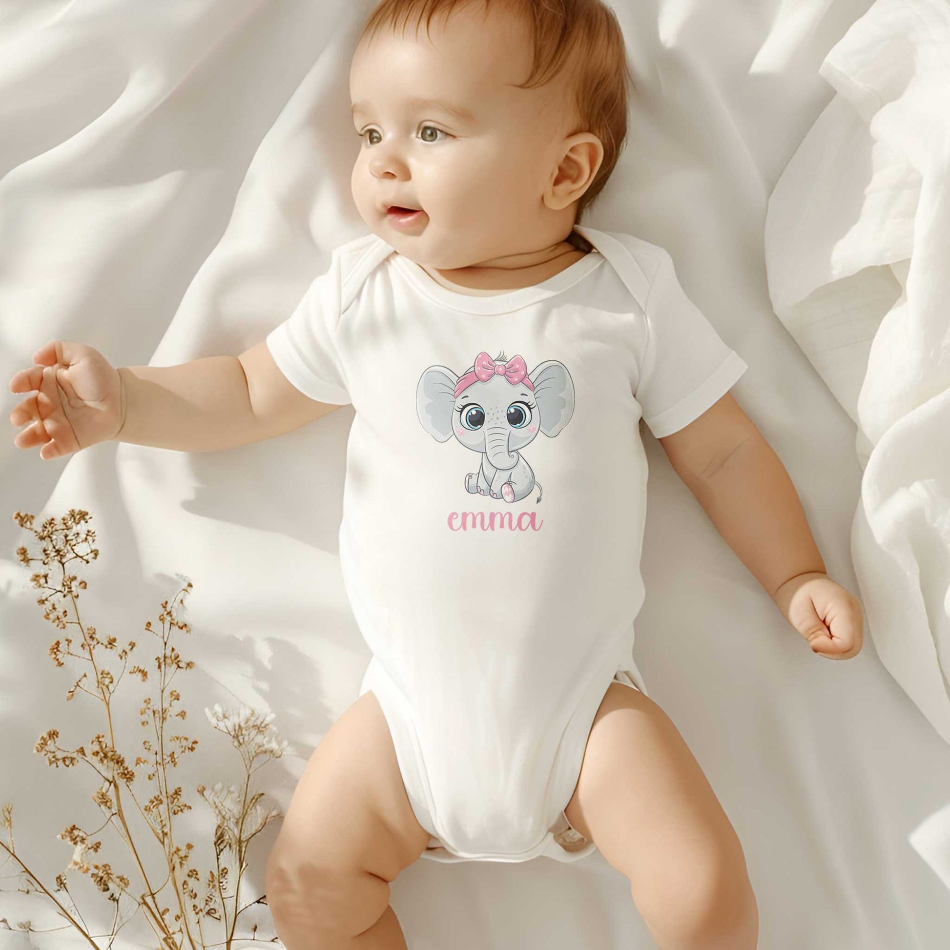 Baby met licht haar ligt op een wit dekentje in een zacht verlichte kamer, draagt een wit rompertje bedrukt met een roze olifantje en een gepersonaliseerde naam in roze.