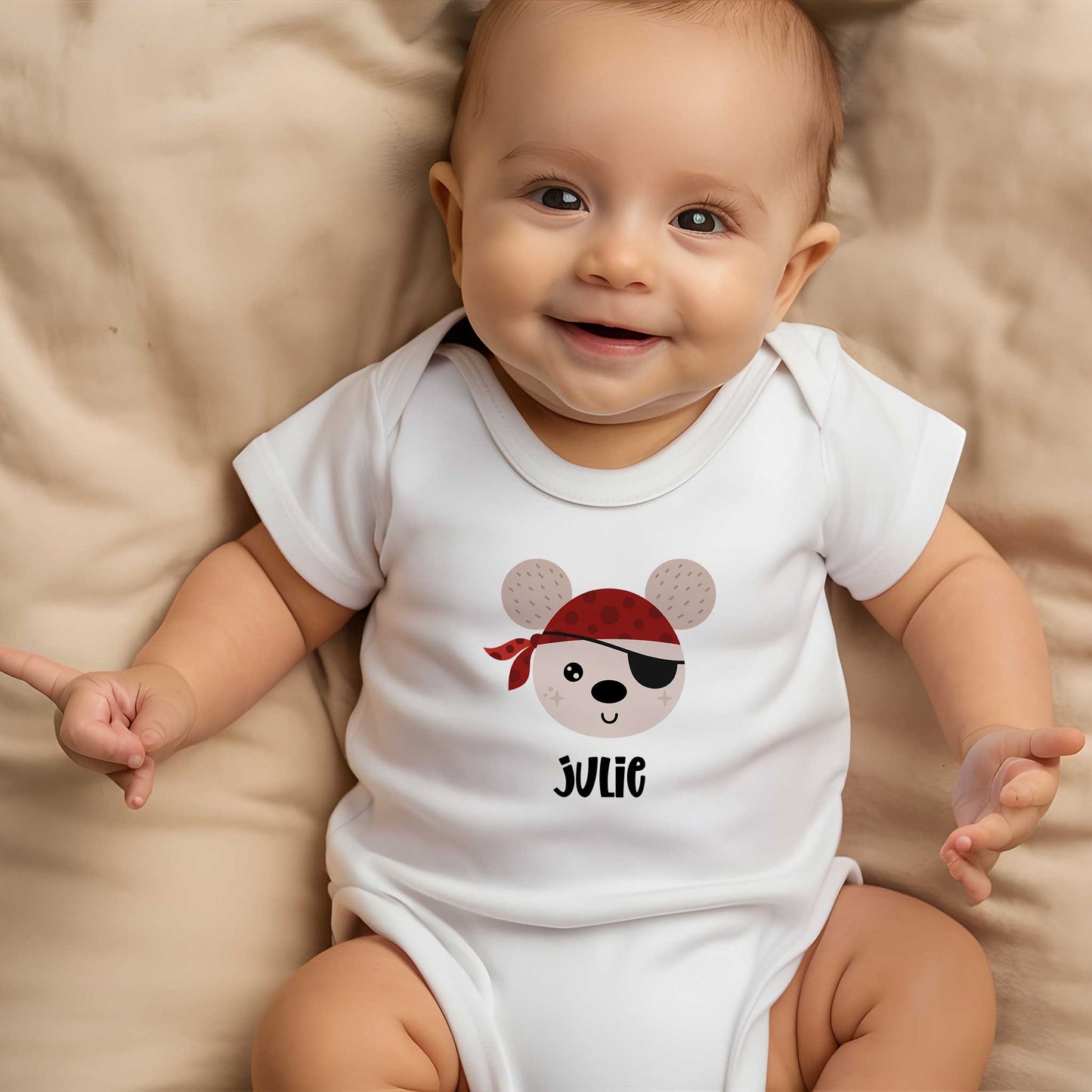 Lachende baby draagt een wit rompertje met een illustratie van een vrolijke piraat en de naam Julie terwijl hij op een zachte beige deken ligt.