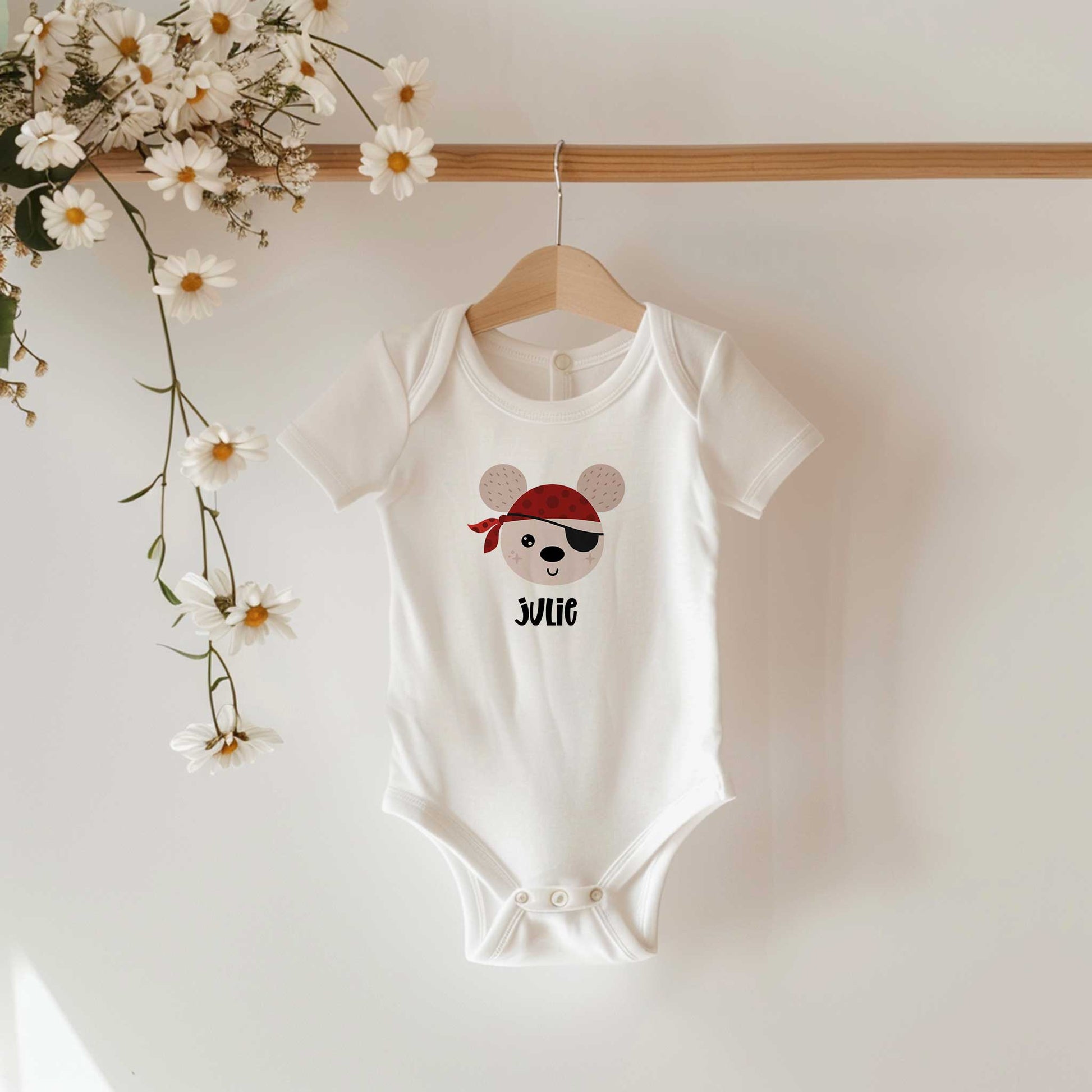 Wit babyrompertje met een illustratie van een vrolijke piraat en de naam Julie, hangend aan een houten kledinghanger met witte madeliefjes op de achtergrond.