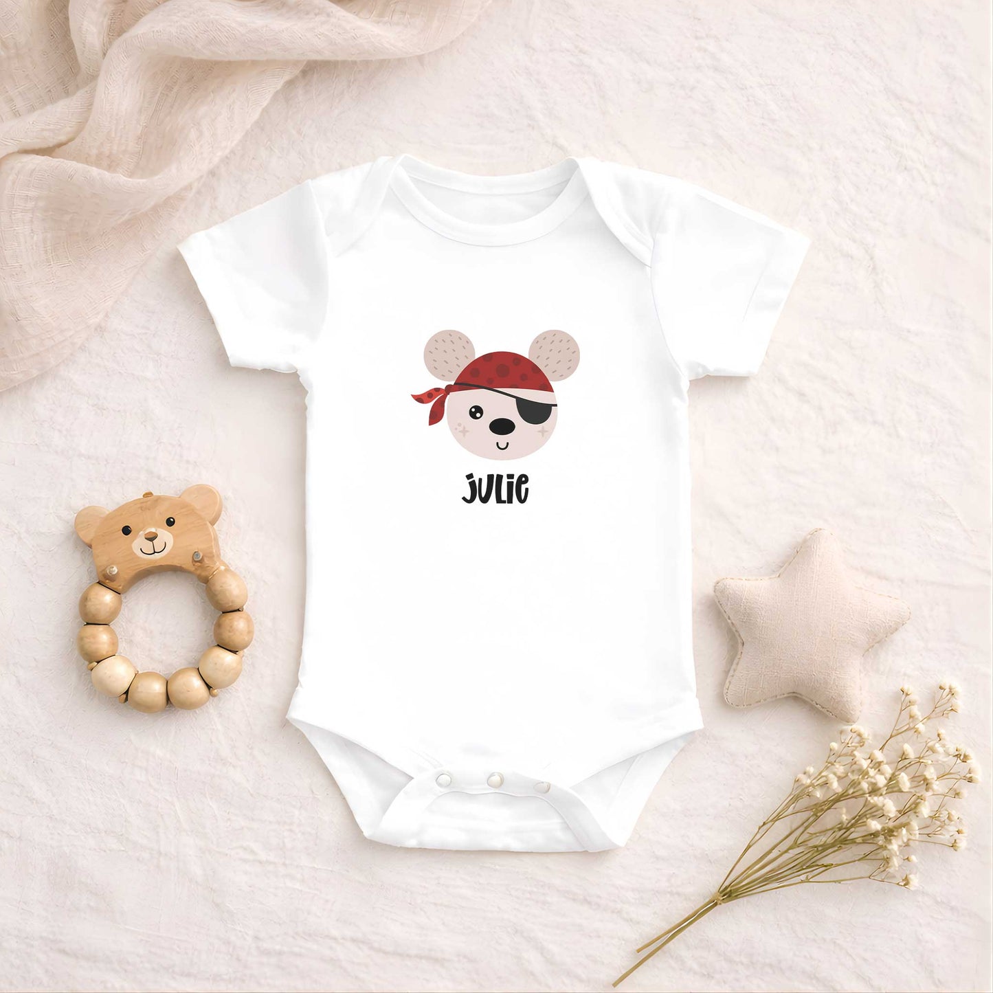 Wit babyrompertje met een illustratie van een vrolijke piraat en de naam Julie, liggend op een zachte beige ondergrond met een houten bijtring, stoffen ster en droogbloemen.