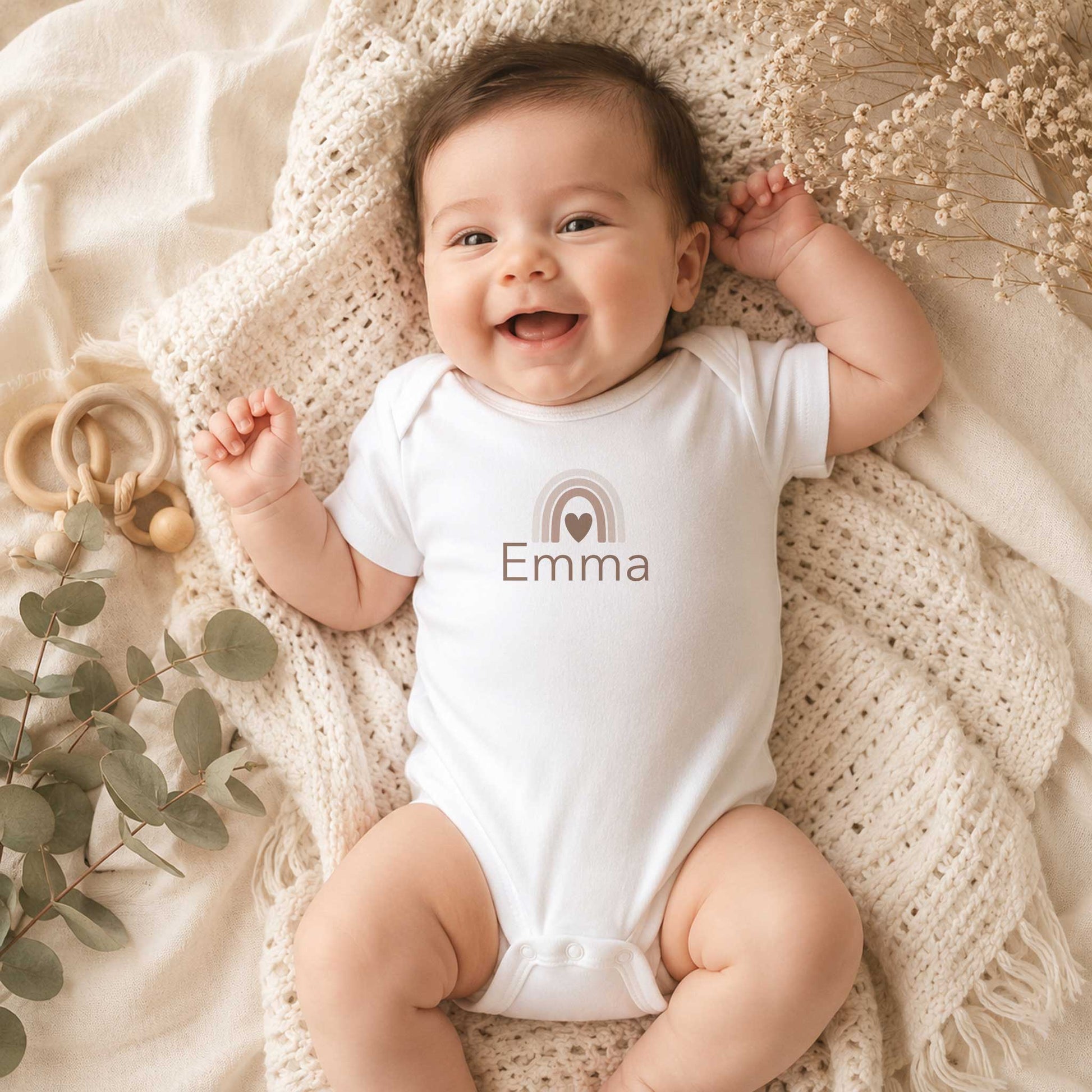 Lachende baby draagt een wit rompertje met een neutrale regenboog en de naam Emma, liggend op een beige gebreide deken.