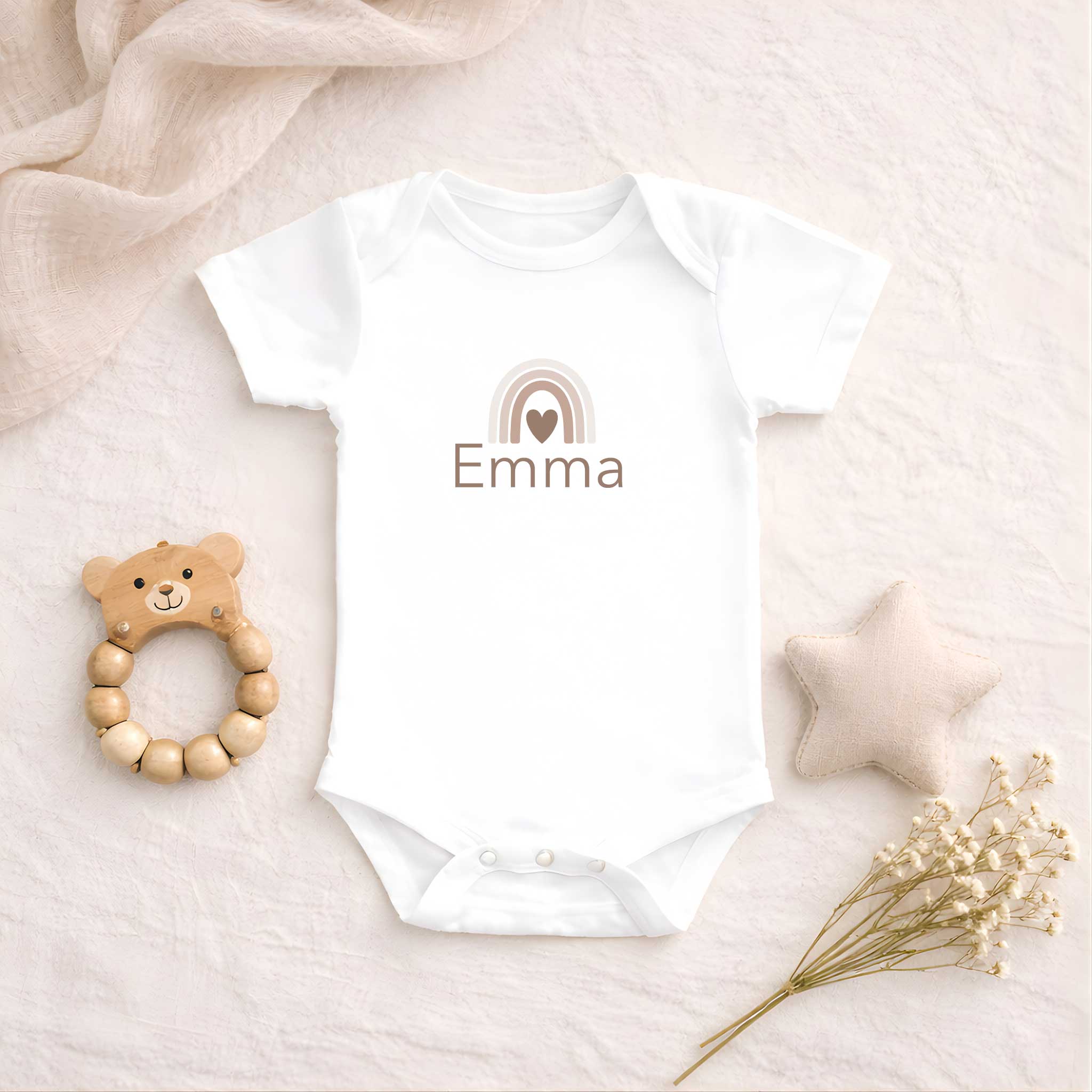 Wit babyrompertje met een neutrale regenboog en de naam Emma, liggend op een zachte beige ondergrond naast een houten bijtring, een stoffen ster en droogbloemen.