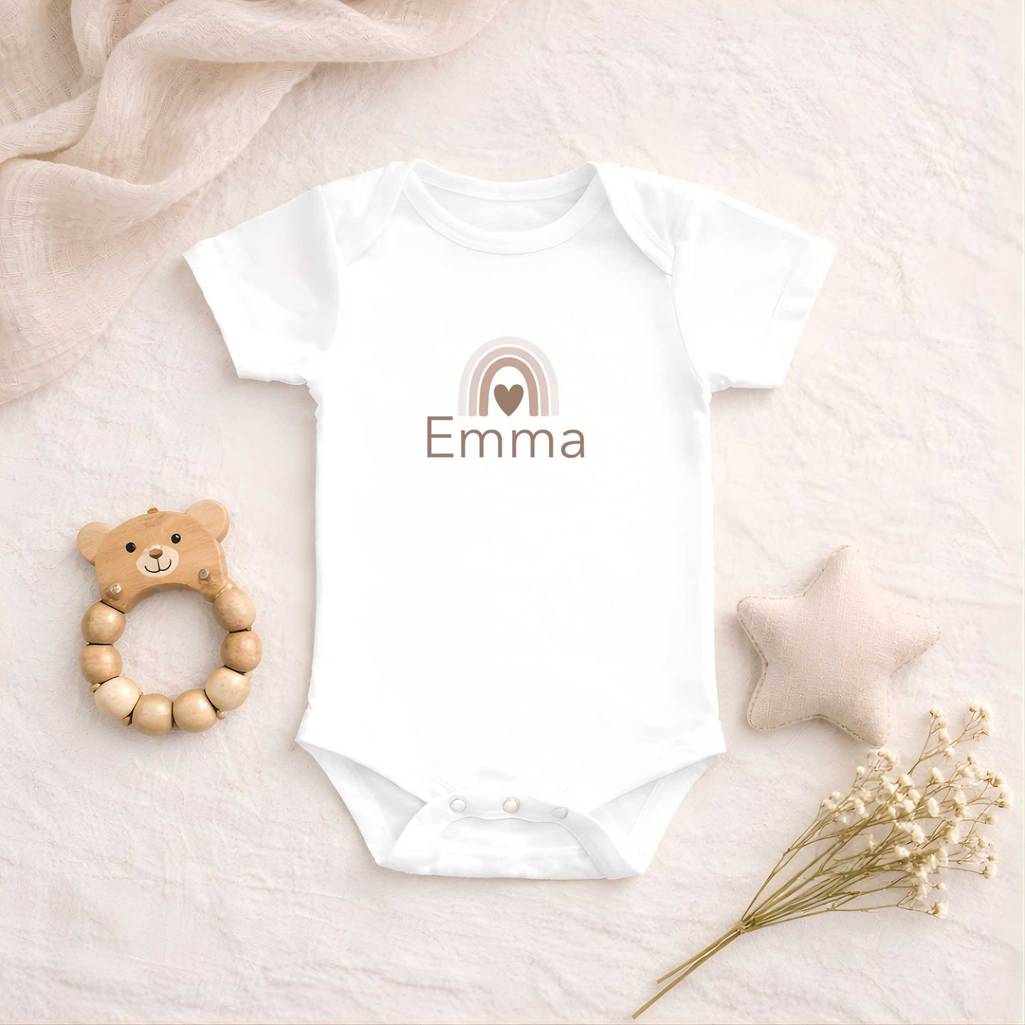 Wit babyrompertje met een neutrale regenboog en de naam Emma, liggend op een zachte beige ondergrond naast een houten bijtring, een stoffen ster en droogbloemen.
