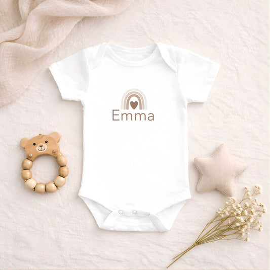 Wit babyrompertje met een neutrale regenboog en de naam Emma, liggend op een zachte beige ondergrond naast een houten bijtring, een stoffen ster en droogbloemen.