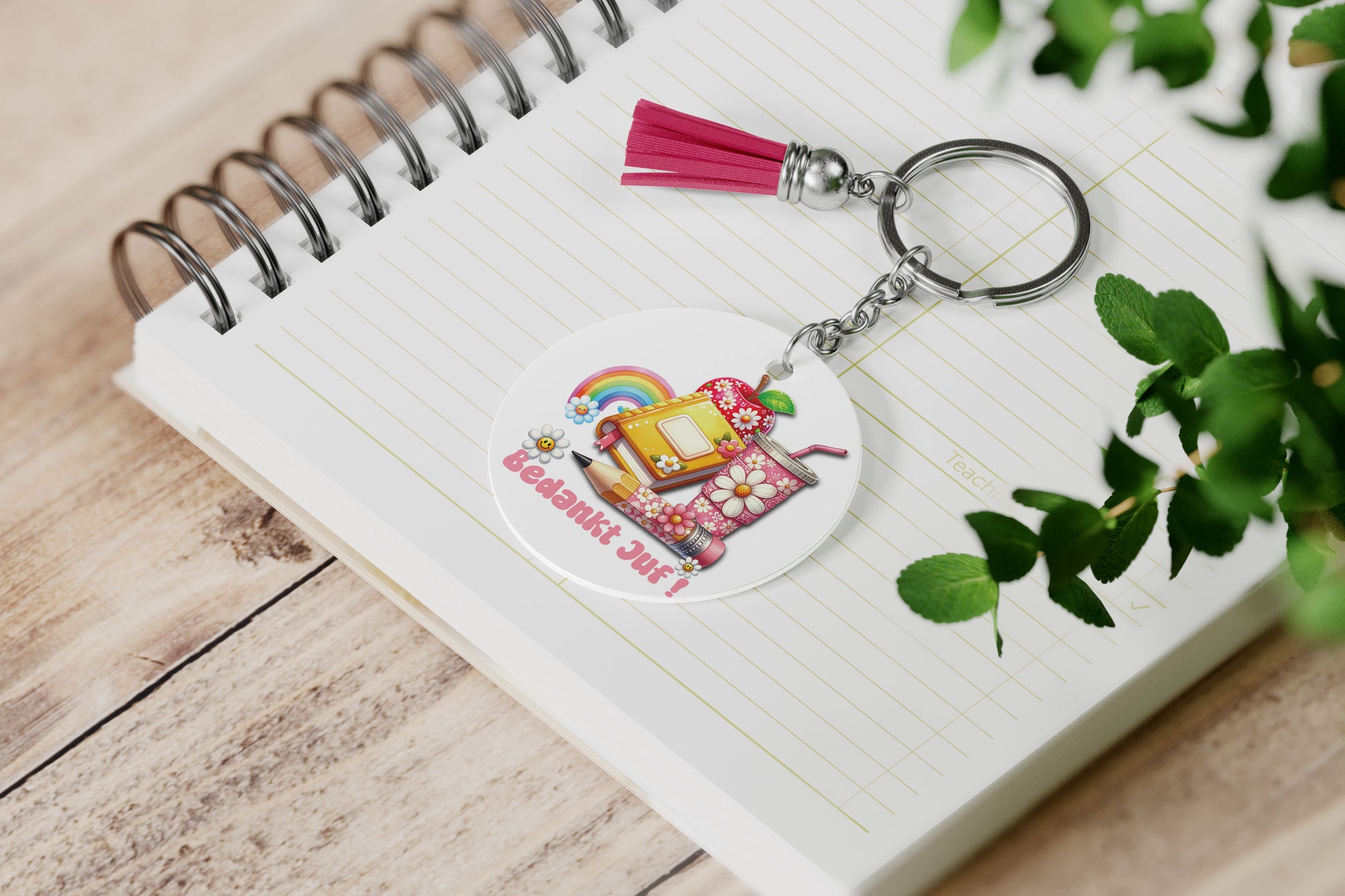 ronde sleutelhanger voor juf met de tekst bedankt juf in roze en groovy illustraties zoals een appel, boek, regenboog en beker met bloemen lggend op een notieboek met een roze kwastje