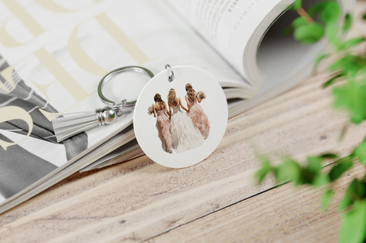 ronde witte sleutelhanger met waterverf illustratie in zachte kleuren van een bruid en bruismeisjes gezien van de achterkant , sleutelhanger heeft wit kwastje. Sleuthanger ligt opeen opgengeslagen magazine.