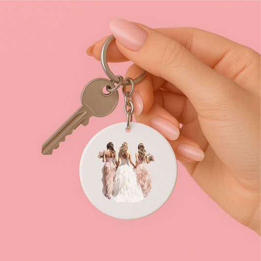 vrouwenhand die een ronde witte sleutelhanger met waterverf illustratie in zachte kleuren van een bruid en bruismeisjes gezien van de achterkant vasthoudt, sleutelhanger heeft wit kwastje