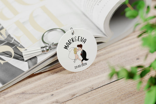 gepersonaliseerde slutelhanger voor bruispaar met namen en trouwdatum en een illustratie in de vorm van een cartoon bruilofststel. Aan de hanger hangt een wit suede kwastje op een tafel liggend tegen een opengeslagen magazine