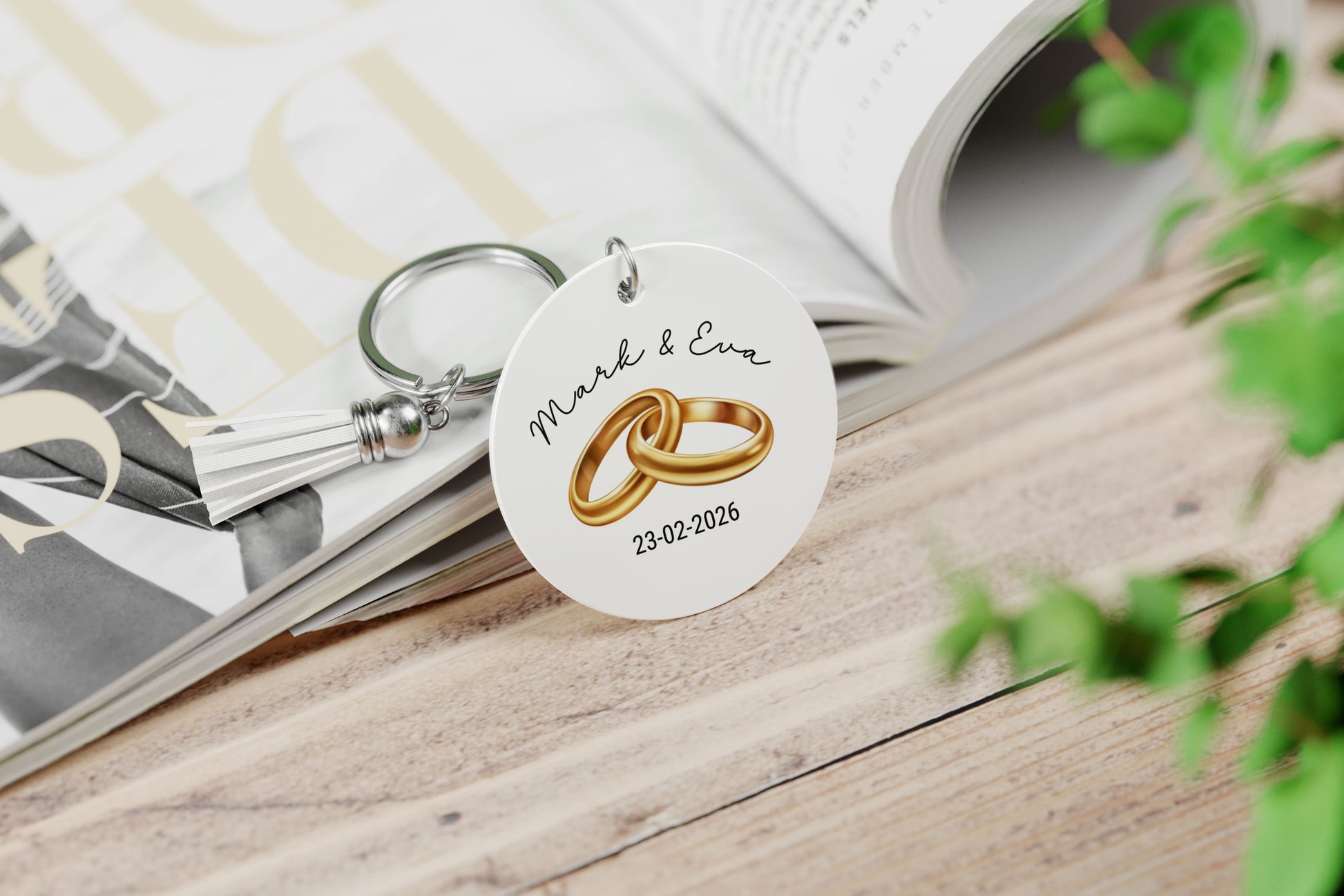 wit ronde sleutelhanger met wit kwastje, met illustratie van 2 gouden trouwringen , namen van het aanstaande echtpaar en hun trouwdatum op een opengeslagen magazine
