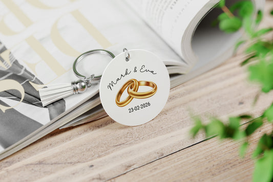 wit ronde sleutelhanger met wit kwastje, met illustratie van 2 gouden trouwringen , namen van het aanstaande echtpaar en hun trouwdatum op een opengeslagen magazine