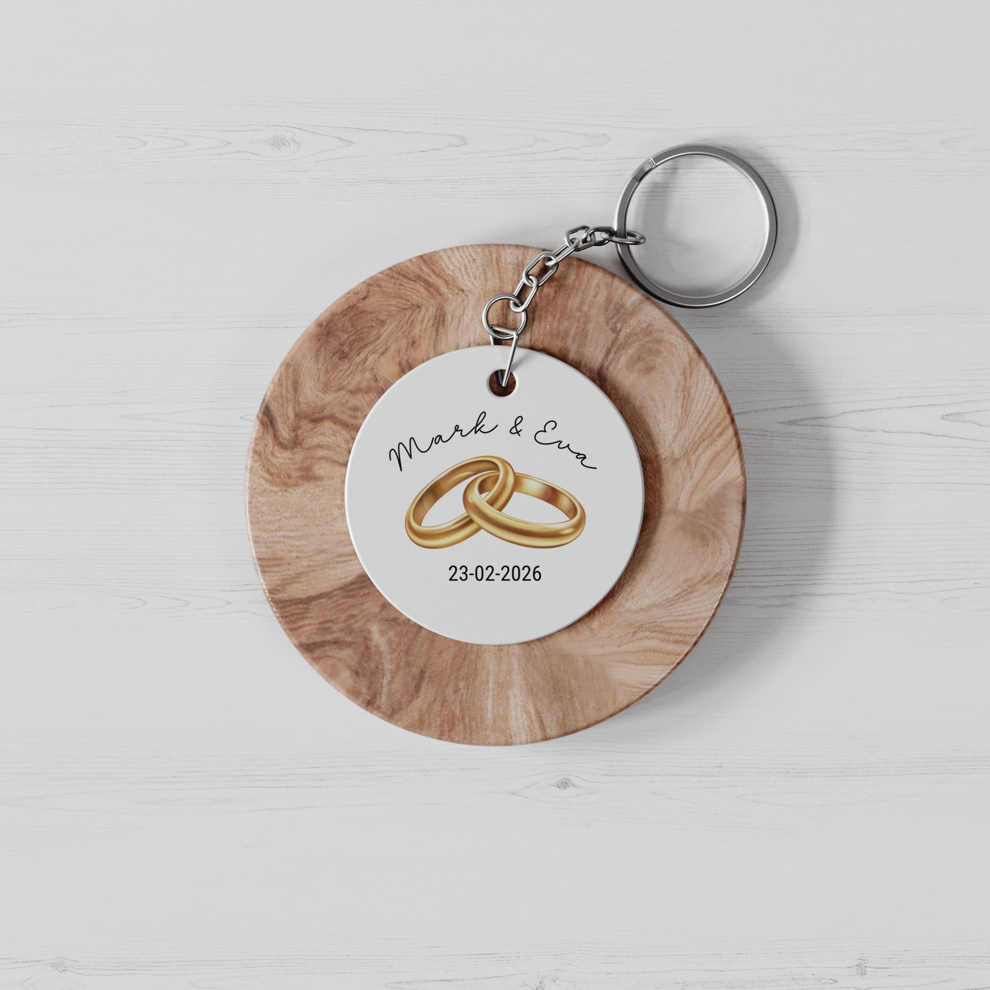 witte ronde sleutelhanger met illustratie van 2 gouden trouwringen , namen van het aanstaande echtpaar en hun trouwdatum op een houten cirkel