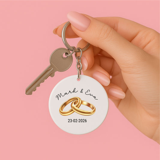 vrouwenhand  met witte ronde sleutelhanger met illustratie van 2 gouden trouwringen , namen van het aanstaande echtpaar en hun trouwdatum, tevens sleutel aan de sleutel ring