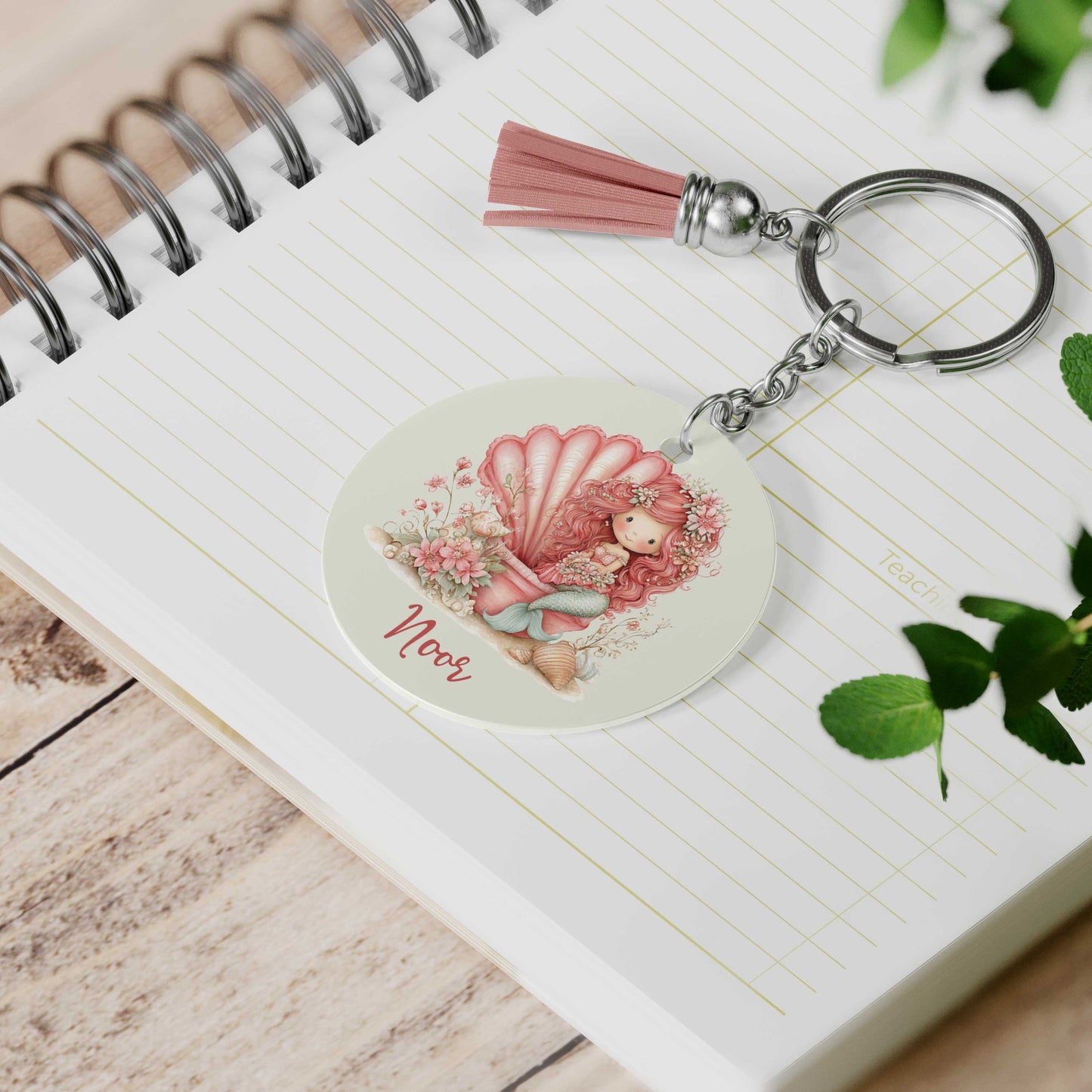 Ronde sleutelhanger met roze kwastje liggend op een notitieboek. De sleutelhanger toont een roze zeemeermin in een grote schelp, omringd door bloemen en schelpen, met de naam Noor onder de illustratie.