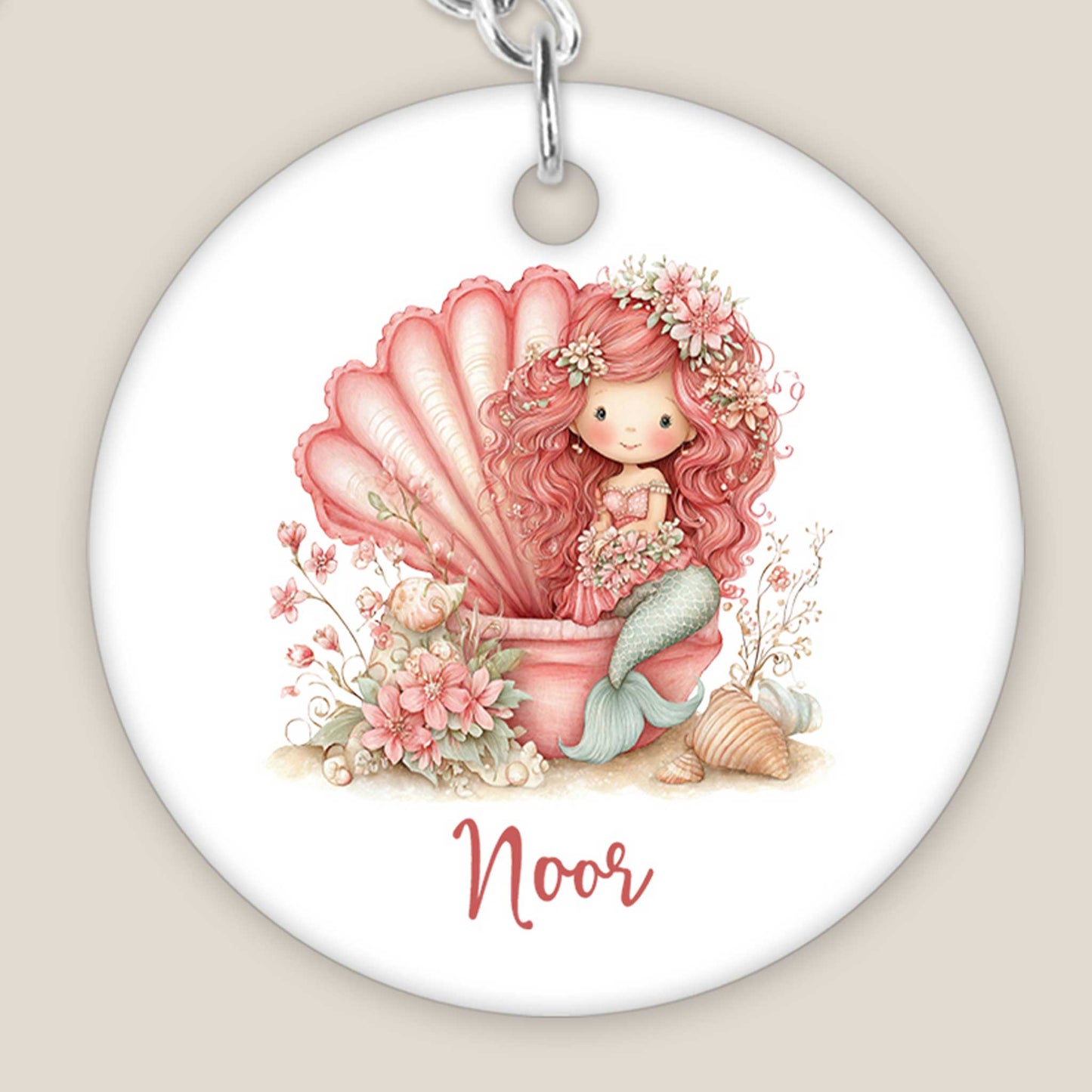 Close-up van een ronde sleutelhanger met een illustratie van een roze zeemeermin in een grote schelp. Onder het ontwerp staat de naam Noor. De bloemen, schelpen en details van de zeemeerminstaart zijn duidelijk zichtbaar.