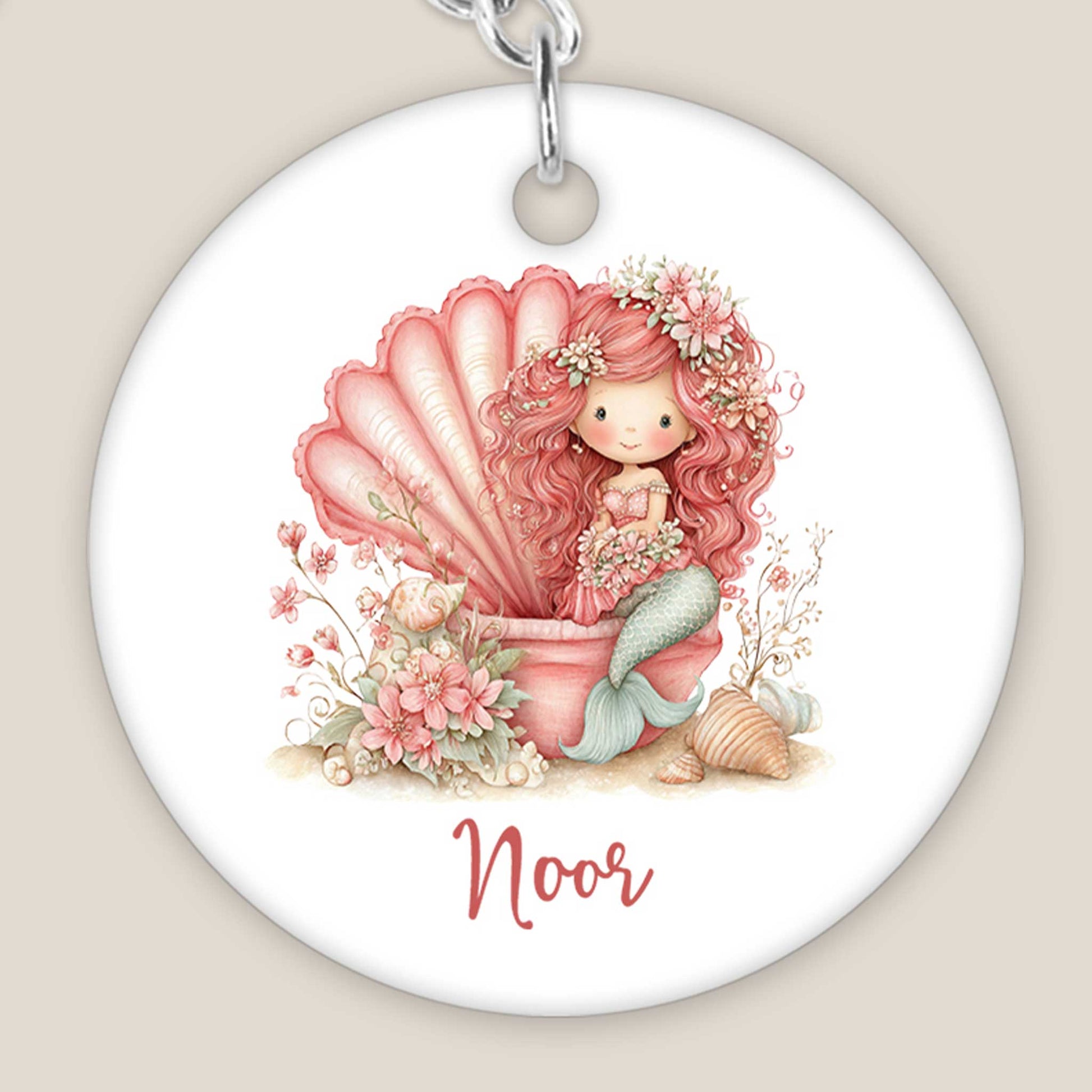 Close-up van een ronde sleutelhanger met een illustratie van een roze zeemeermin in een grote schelp. Onder het ontwerp staat de naam Noor. De bloemen, schelpen en details van de zeemeerminstaart zijn duidelijk zichtbaar.