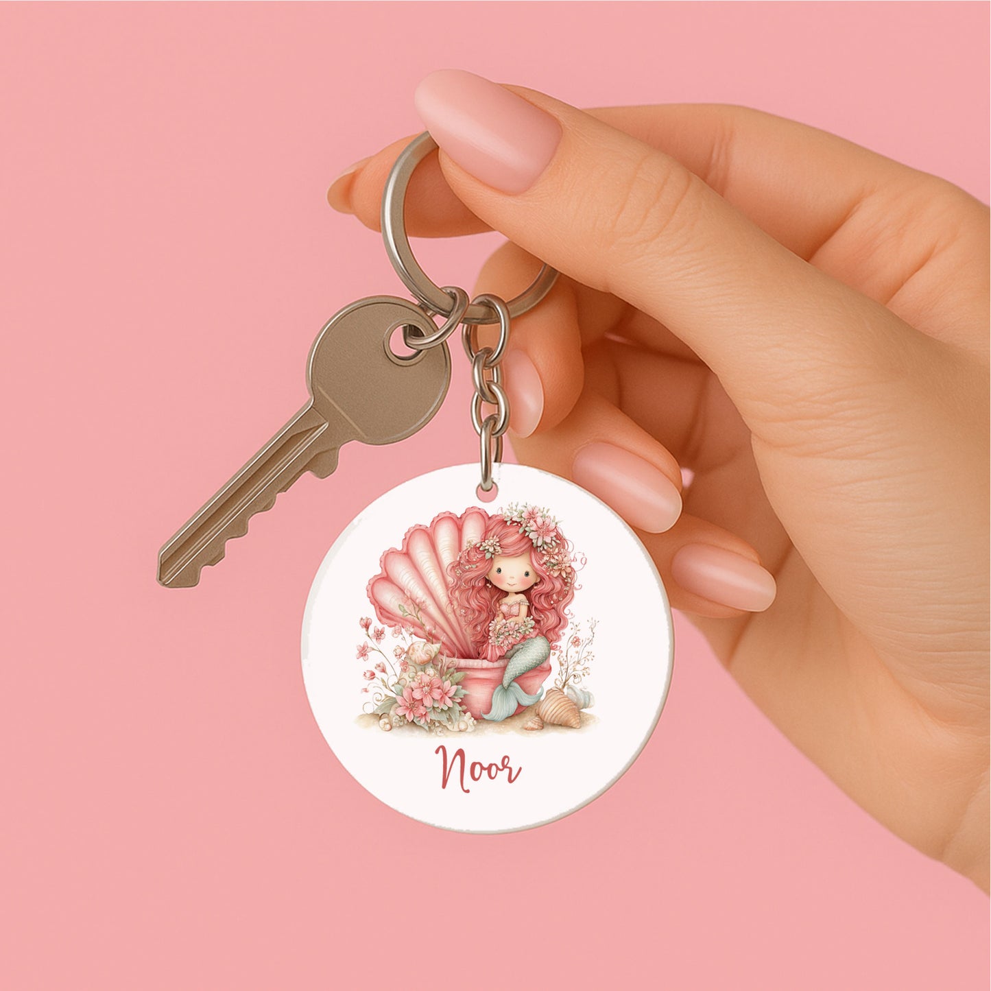 Hand houdt een gepersonaliseerde ronde sleutelhanger vast aan een sleutelring. Op de sleutelhanger staat een roze zeemeermin in een schelp met bloemen, schelpen en de naam Noor tegen een roze achtergrond.