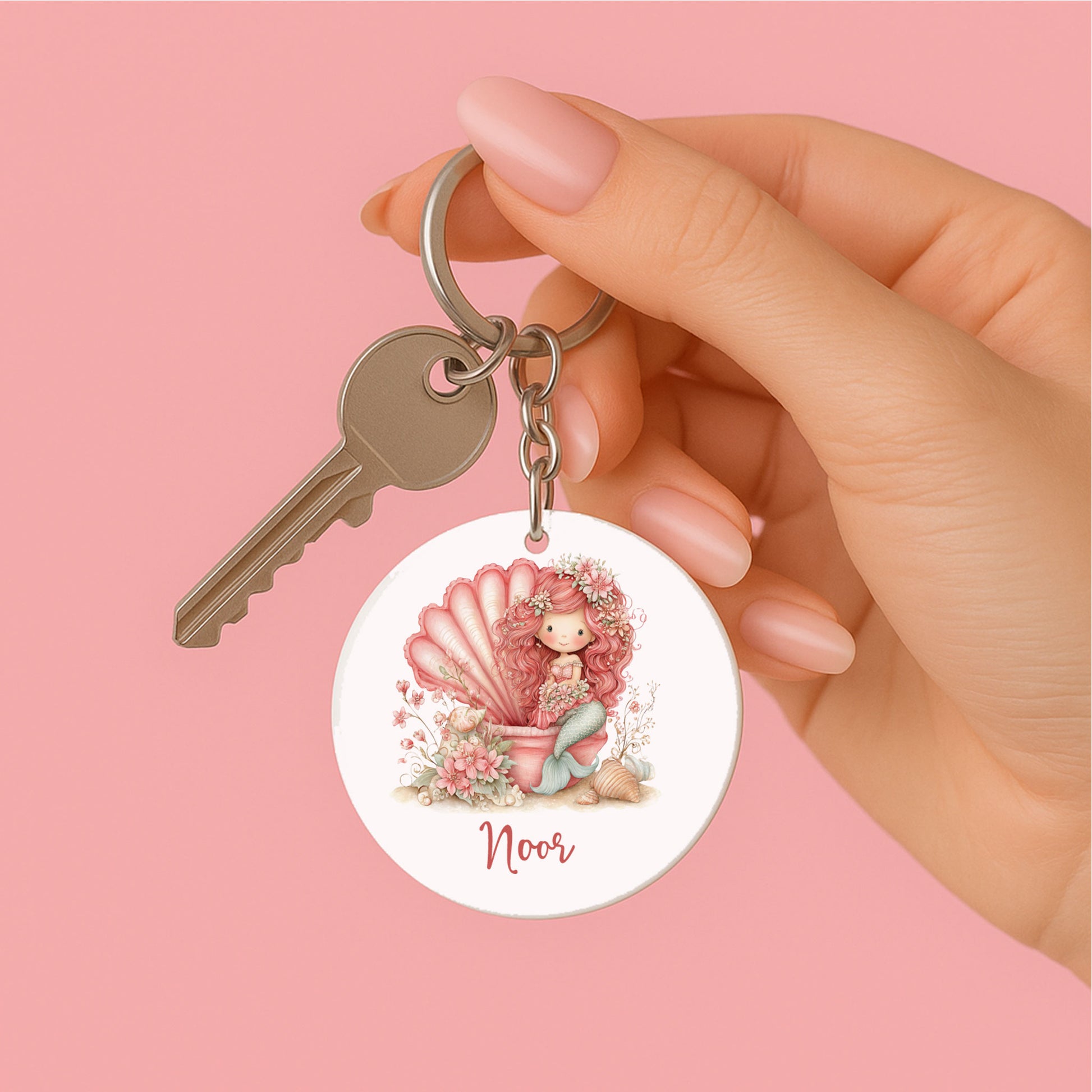 Hand houdt een gepersonaliseerde ronde sleutelhanger vast aan een sleutelring. Op de sleutelhanger staat een roze zeemeermin in een schelp met bloemen, schelpen en de naam Noor tegen een roze achtergrond.