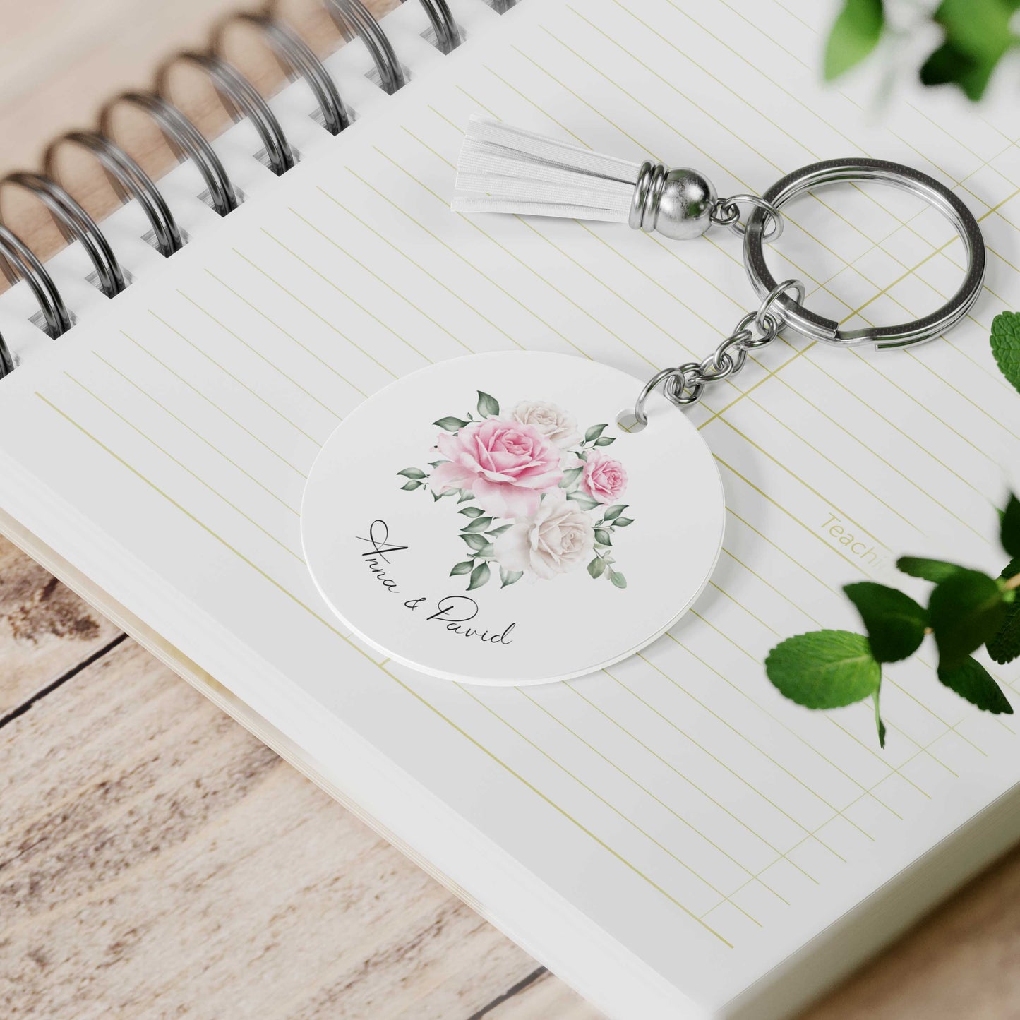 Ronde sleutelhanger met bloemenillustratie, twee namen, witte kwast en metalen sleutelring ligt op een open notitieboek.