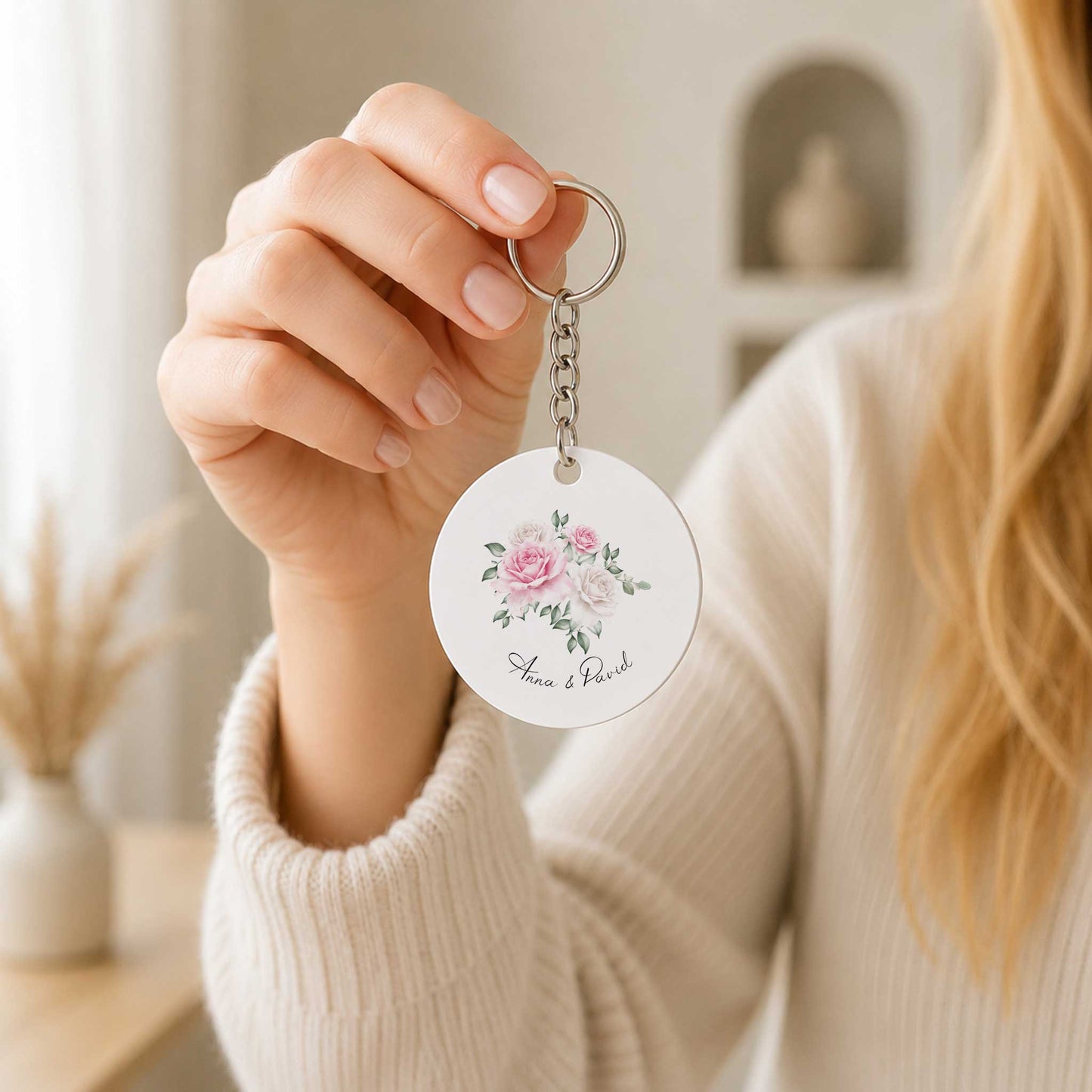Vrouw in een lichte trui houdt een ronde sleutelhanger met roze en witte bloemenillustratie en de namen van het bruidspaar vast.