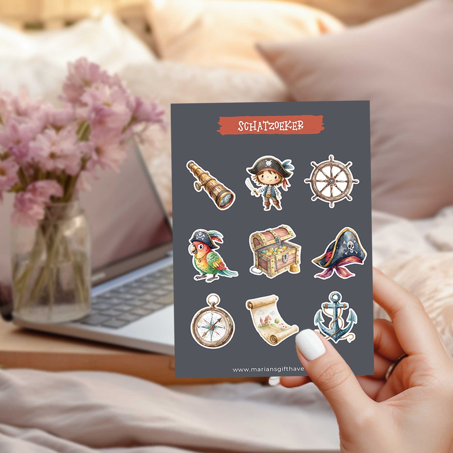 Sfeerfoto van een hand die een stickervel met negen piratenstickers vasthoudt met een laptop en bloemen op de achtergrond.