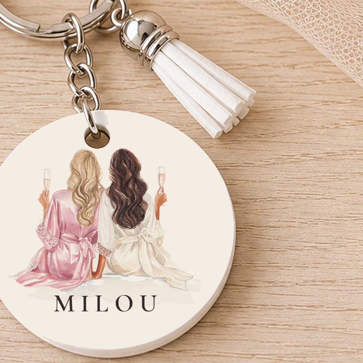 Close-up van een ronde sleutelhanger met twee vrouwen in roze en witte kleding, champagneglazen en de naam Milou