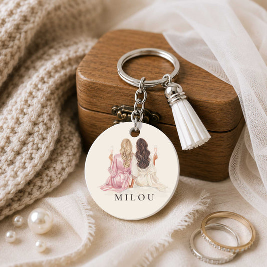 Ronde sleutelhanger met illustratie van twee vrouwen met champagneglazen en de naam Milou, liggend bij een houten kistje