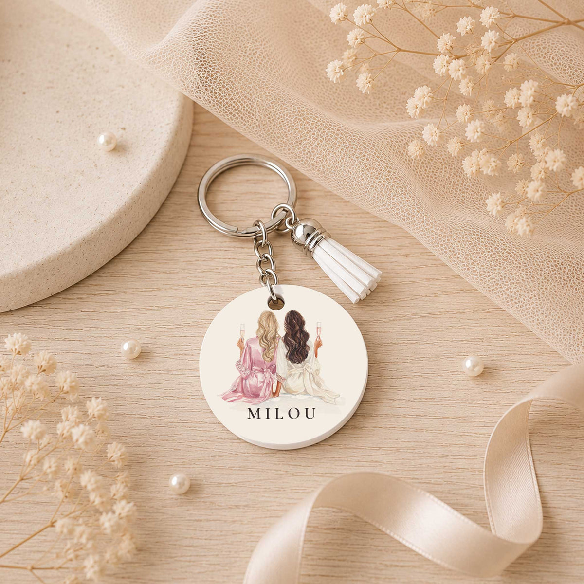 Ronde sleutelhanger met twee vrouwen met champagneglazen en de naam Milou, liggend tussen lint, parels en droogbloemen