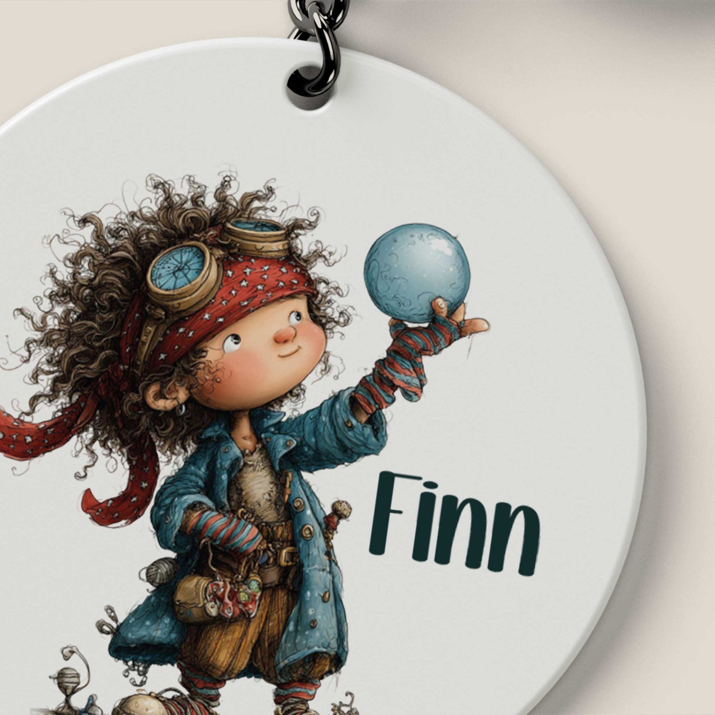 Close-up van een ronde witte sleutelhanger met een illustratie van een lachende piraat met bruine krullen, rode hoofdband, pilotenbril, een blauw kompas en de naam Finn.