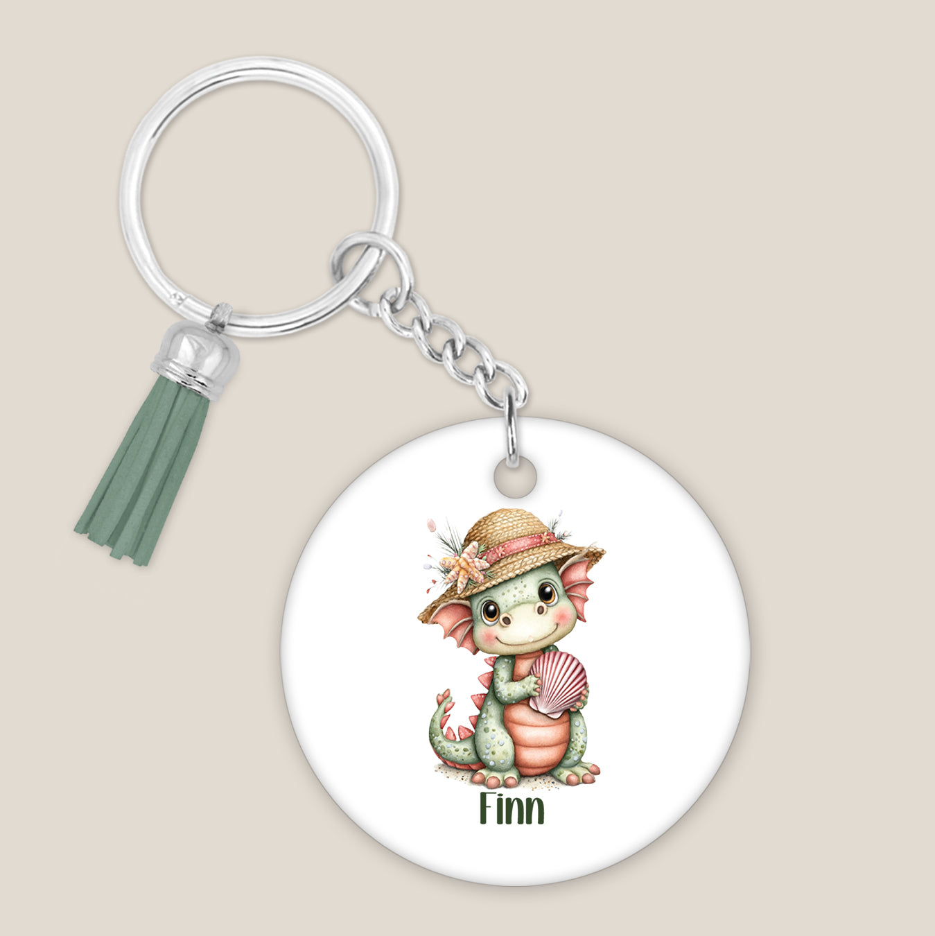 Ronde witte sleutelhanger met een groen draakje met strohoed, roze schelp, de naam Finn en een groene kwast op een beige achtergrond.