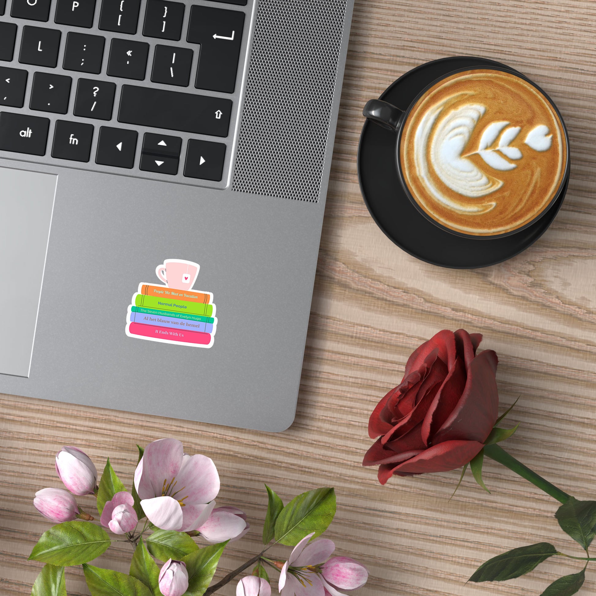 Sticker met boekenstapeldesign op een laptop naast koffie en bloemen, in een gezellige setting.