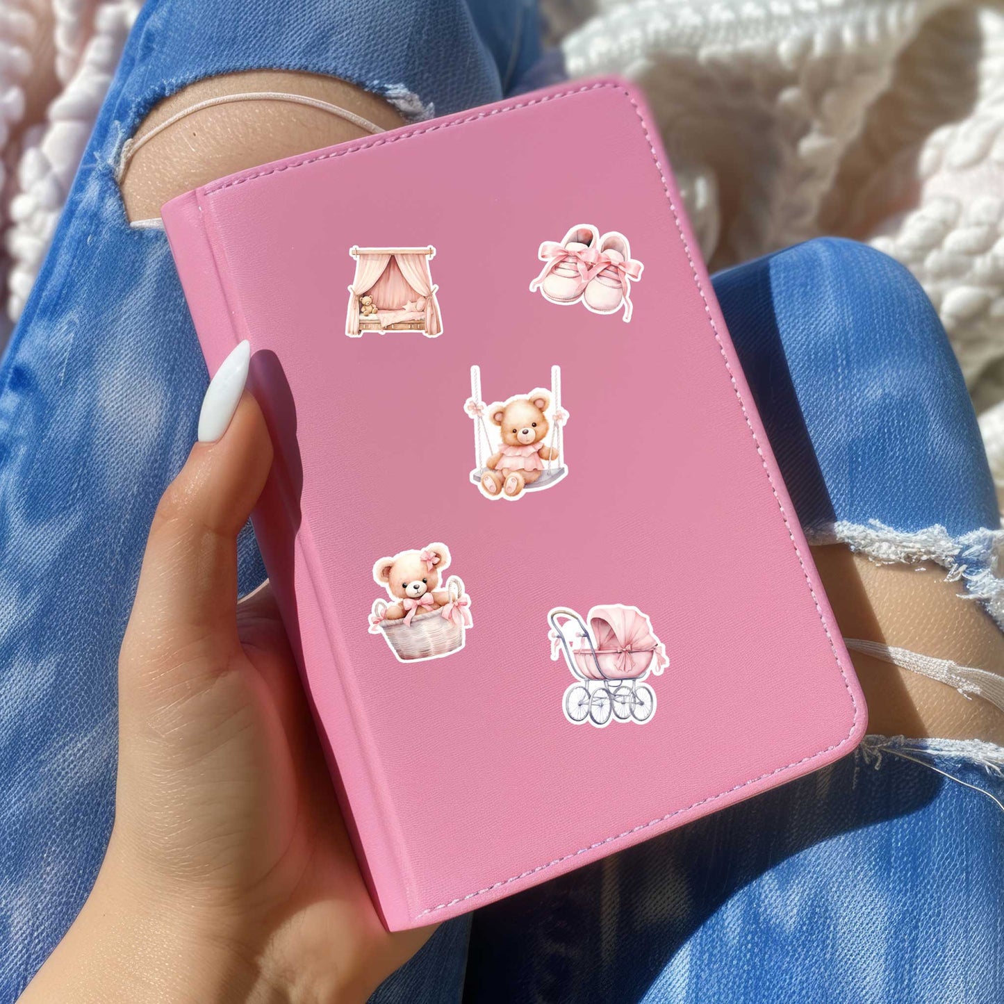 Roze notitieboek versierd met Teddy Baby stickers van een teddybeer op een schommel, teddybeer in een mandje, roze kinderwagen, babyschoentjes en een wiegje met hemel, vastgehouden op schoot.