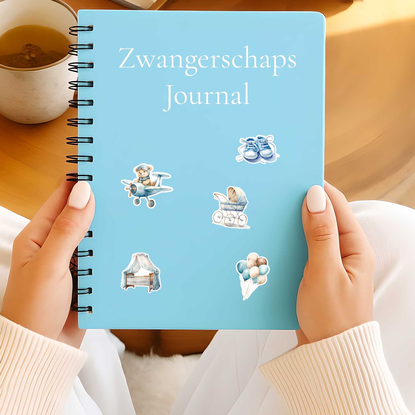 Lichtblauw zwangerschapsjournal met teddy dreams stickers van een teddybeer in een vliegtuigje, kinderwagen, wiegje met hemel, babyschoentjes en ballonnen, vastgehouden door twee handen.